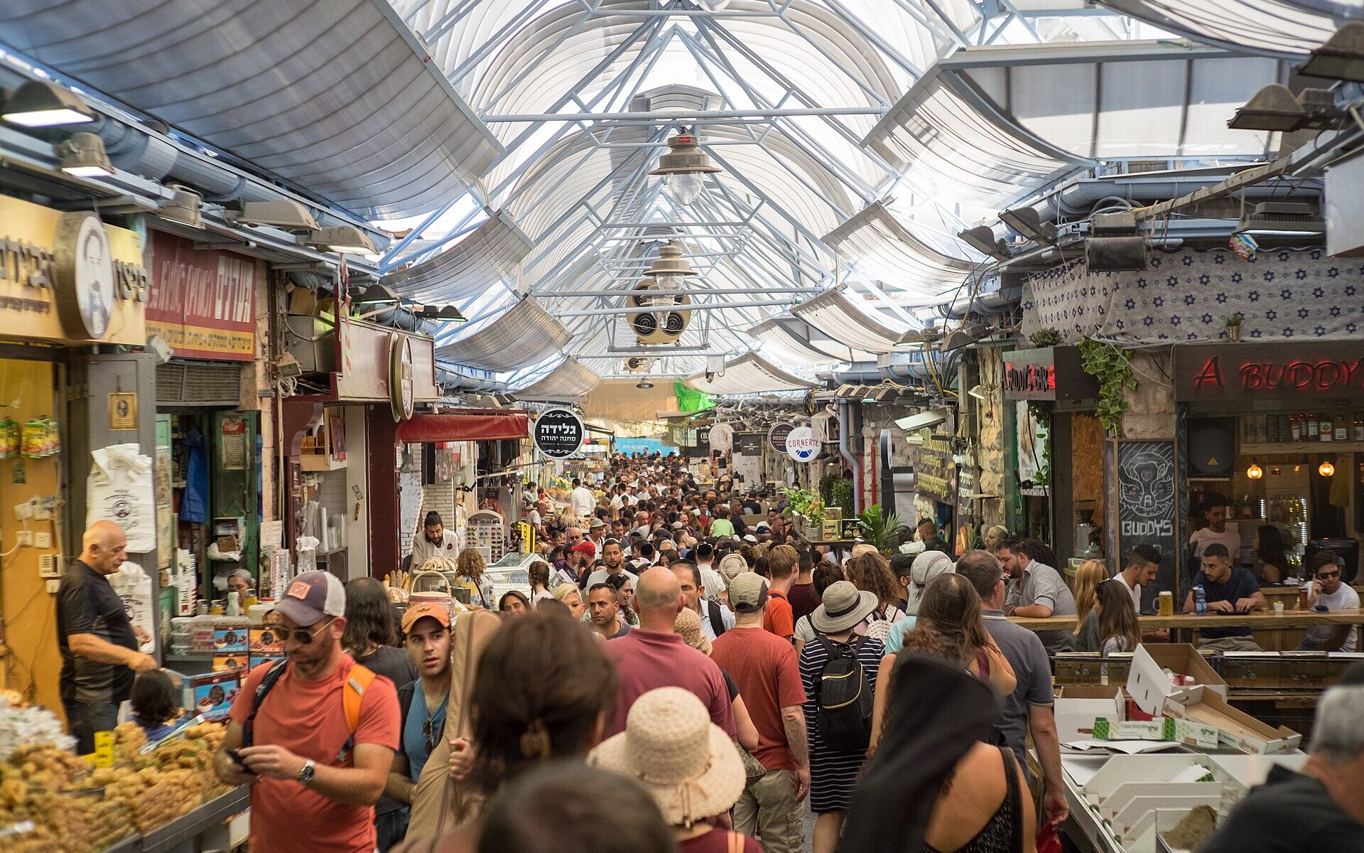 Crowds of shoppers fill a covered lane of Mahane Yehuda Market in Jerusalem, lined with pastry stalls, Hebrew shop signs and a busy bar under the arched roof