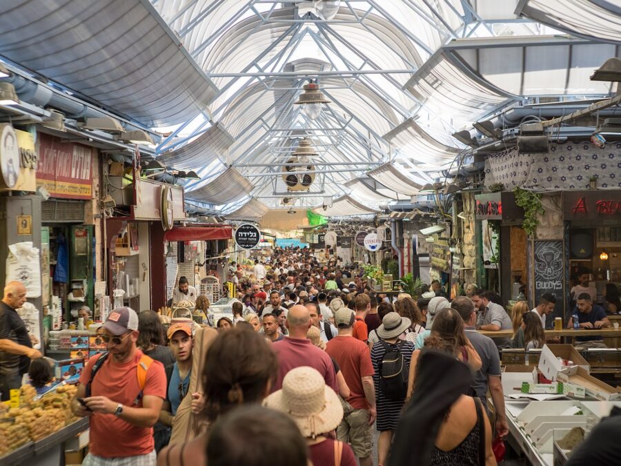 Crowds of shoppers fill a covered lane of Mahane Yehuda Market in Jerusalem, lined with pastry stalls, Hebrew shop signs and a busy bar under the arched roof