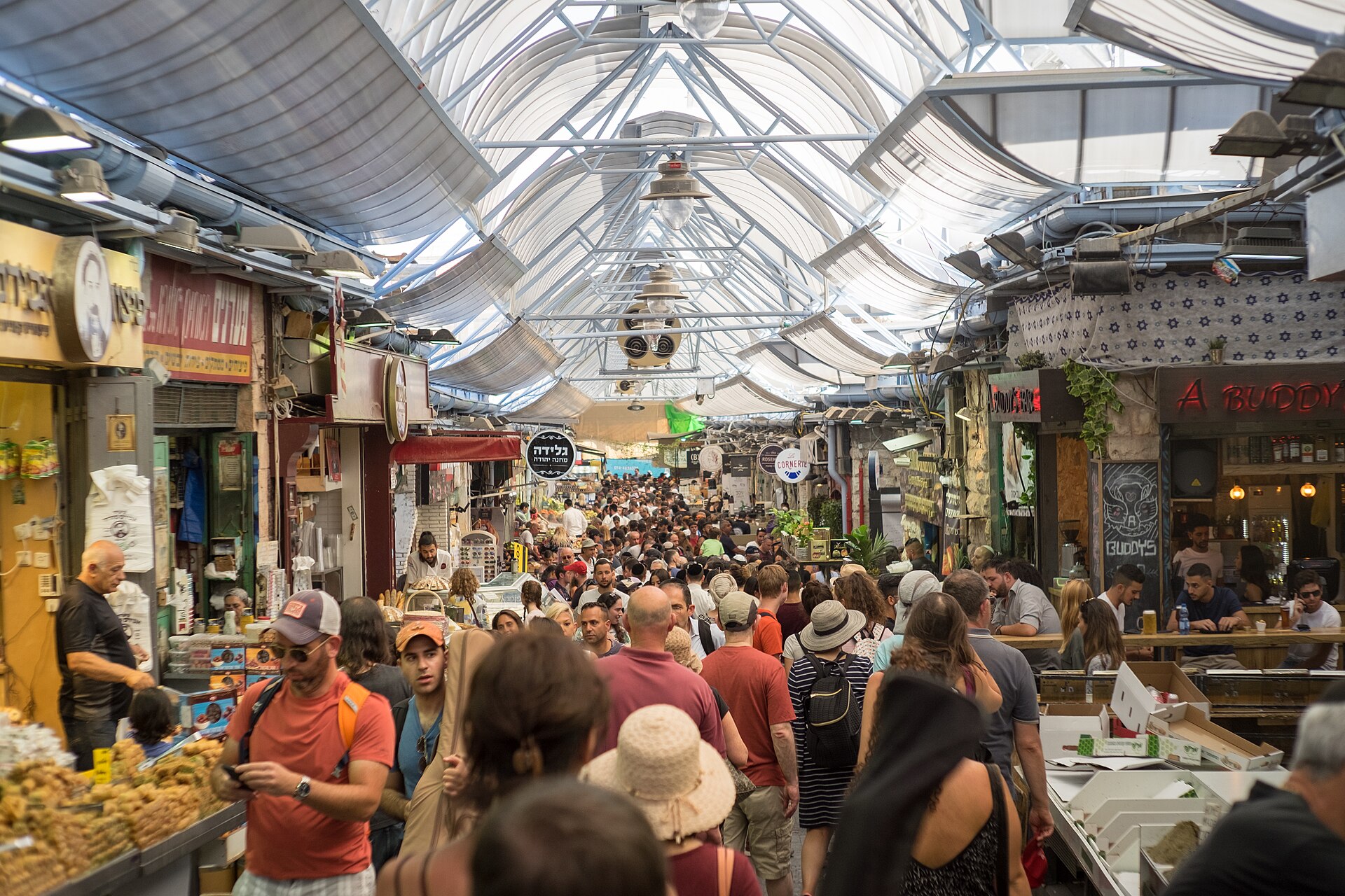 Crowds of shoppers fill a covered lane of Mahane Yehuda Market in Jerusalem, lined with pastry stalls, Hebrew shop signs and a busy bar under the arched roof