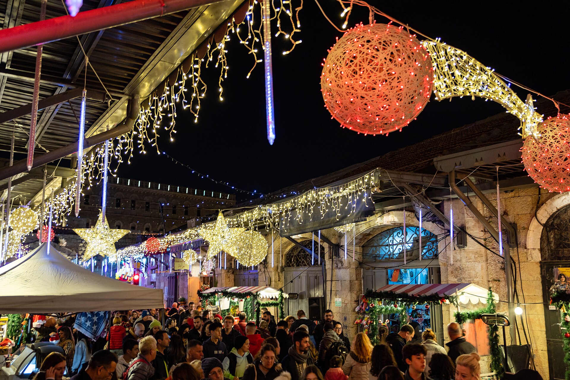 Crowds walk beneath strings of Christmas lights, glowing stars and red baubles at a Christmas market just inside New Gate in Jerusalem's Old City at night