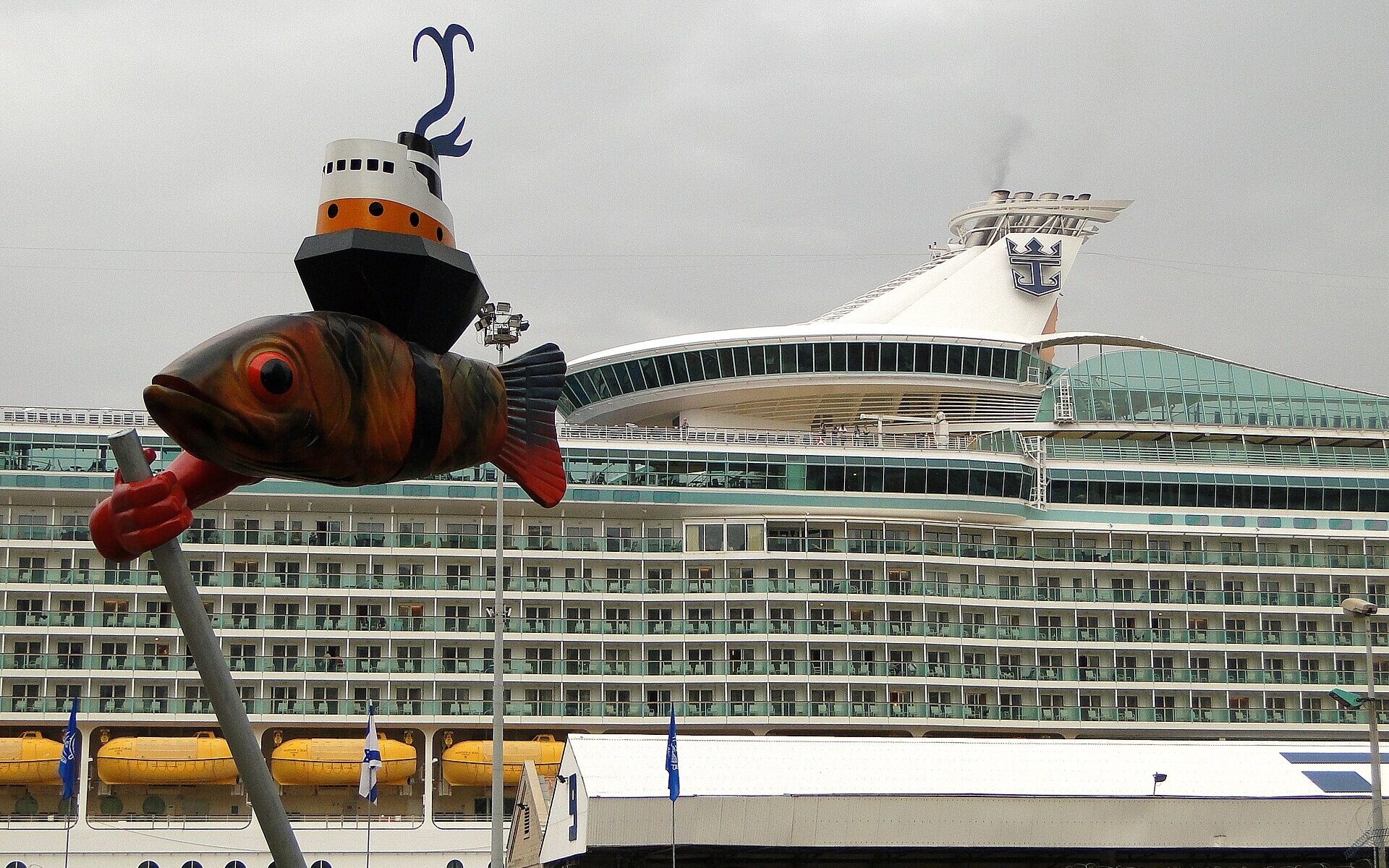 A large Royal Caribbean cruise ship berthed at Haifa Port behind the passenger terminal building, with a whimsical fish-and-boat sculpture in the foreground