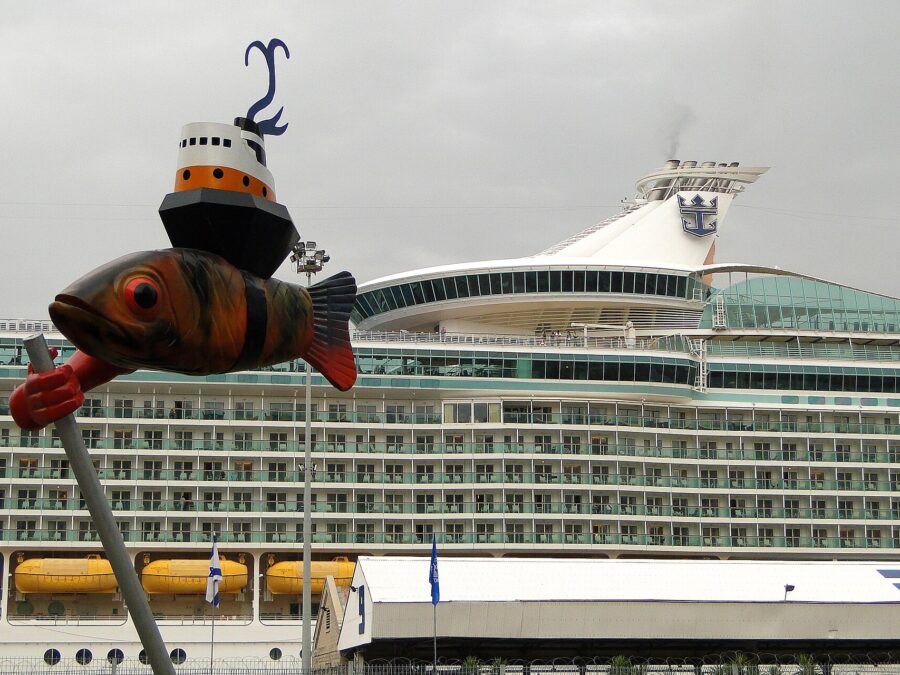 A large Royal Caribbean cruise ship berthed at Haifa Port behind the passenger terminal building, with a whimsical fish-and-boat sculpture in the foreground