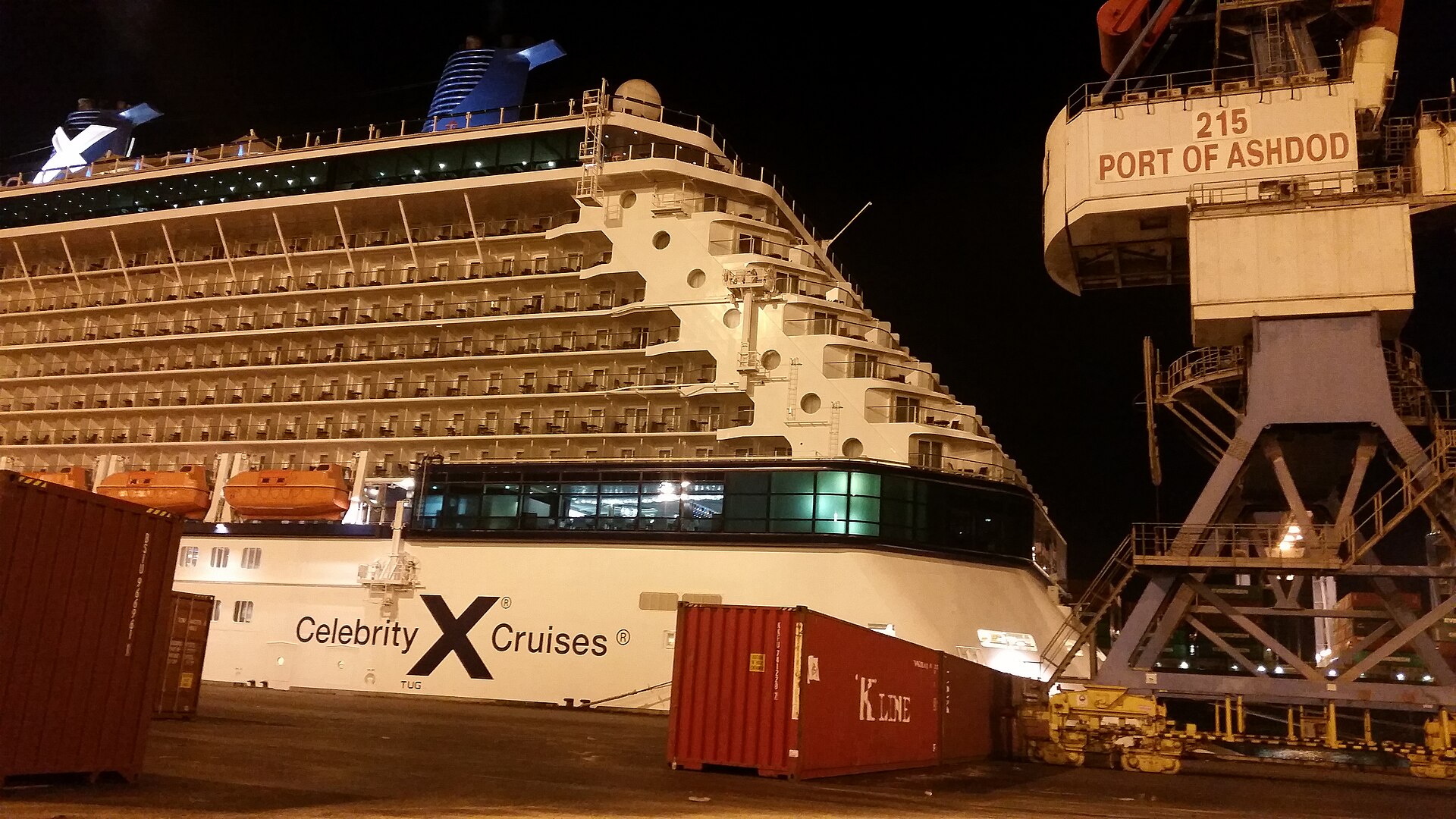A Celebrity Cruises ship berthed at night at the Port of Ashdod, beside a container crane labelled Port of Ashdod and shipping containers on the quay