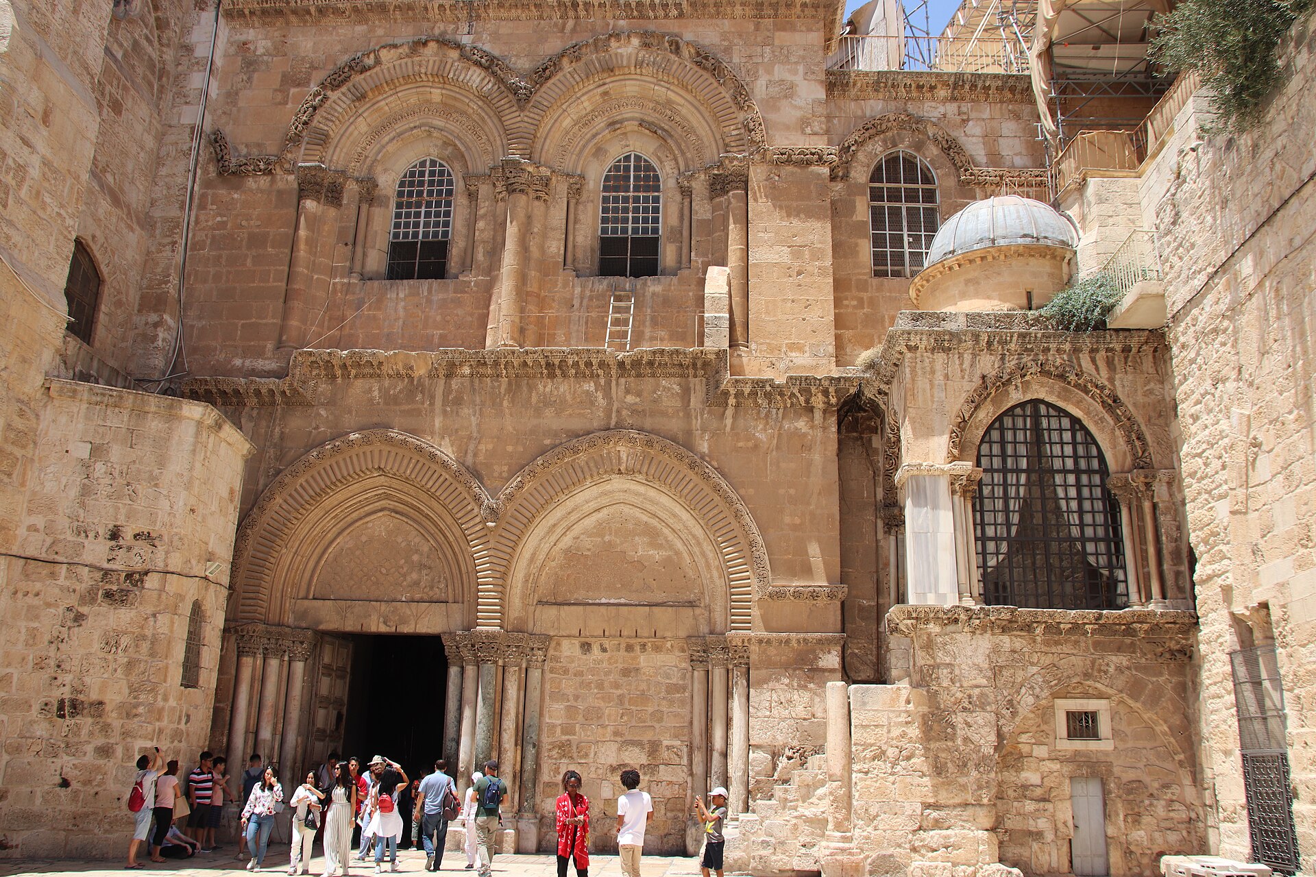 Crusader facade of the Church of the Holy Sepulchre with twin arched doorways, the Immovable Ladder under an upper window and summer visitors at the open door