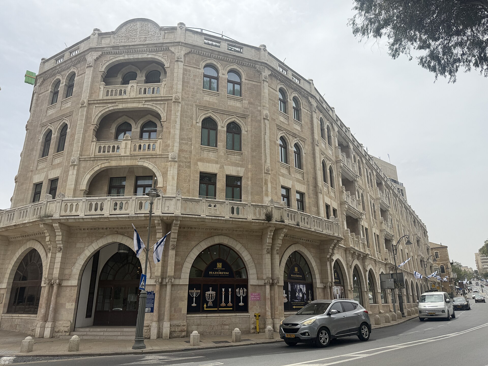 Curved limestone corner façade of the Waldorf Astoria Jerusalem, with Moorish arched windows, stone balconies, an Arabic calligraphy parapet and ground-floor arcades along the street