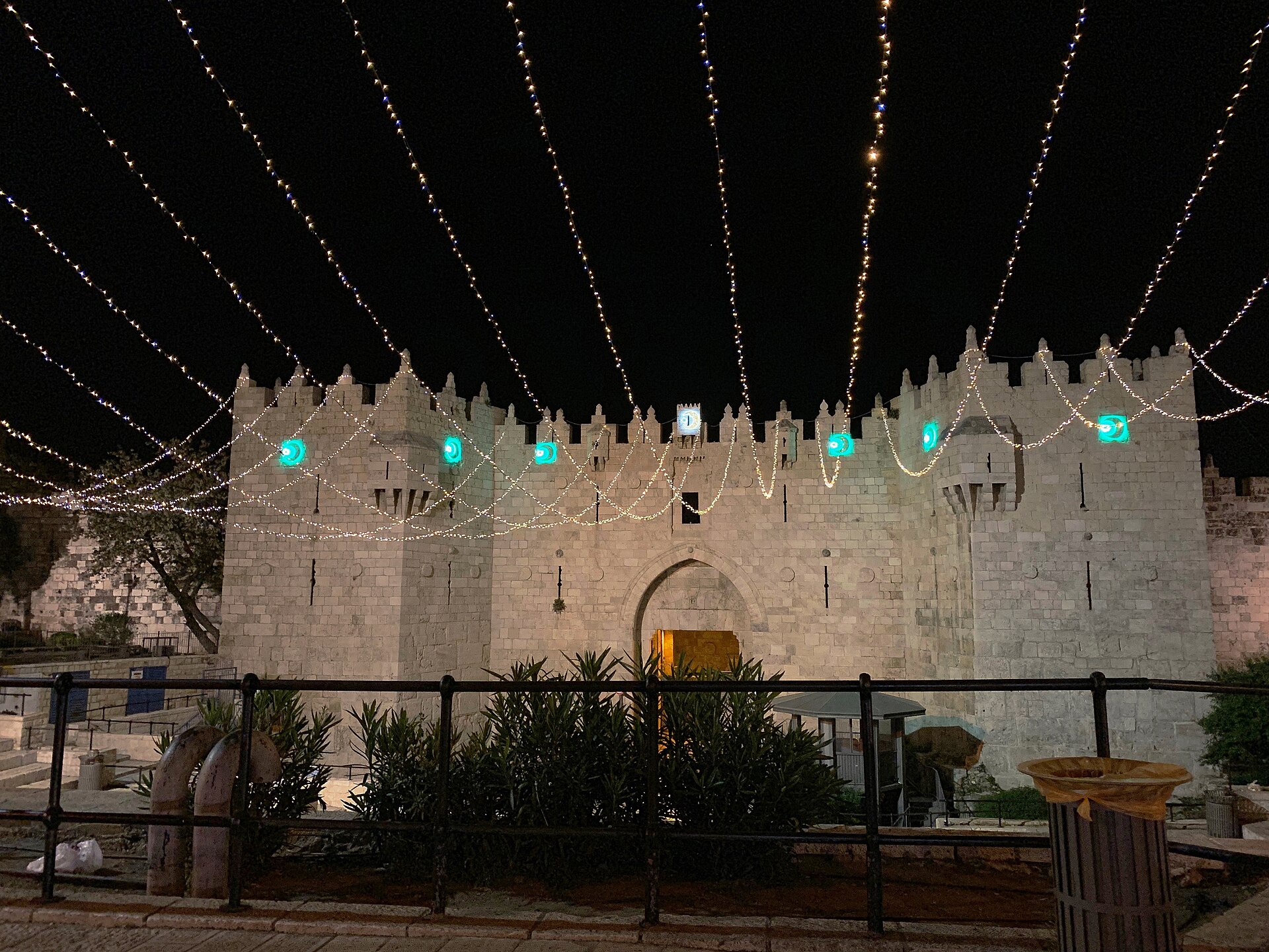 Damascus Gate floodlit at night, with strings of white fairy lights and glowing green crescent-moon lanterns stretched across its crenellated facade and the plaza