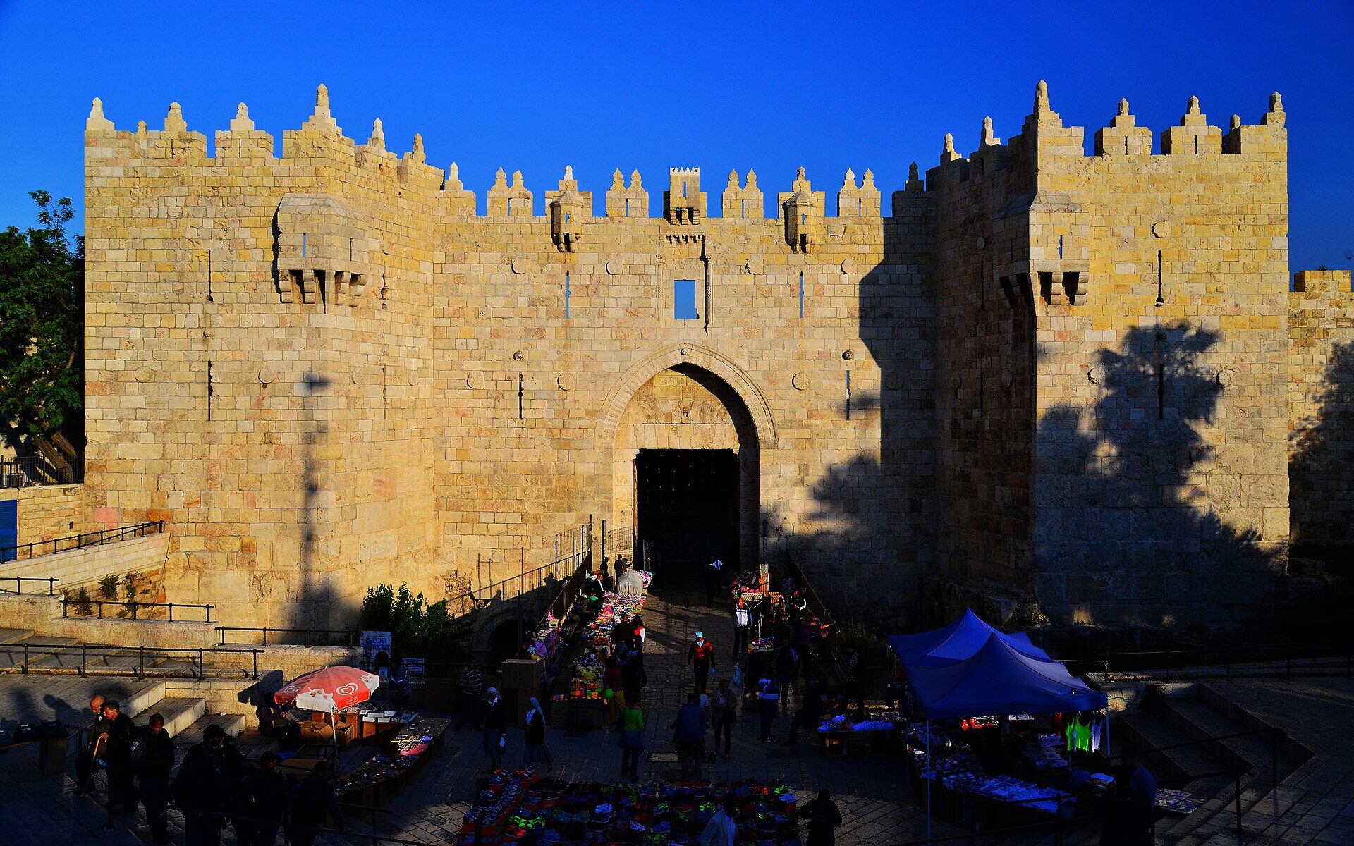 Damascus Gate in Jerusalem's northern Old City wall glowing gold in late-afternoon sun, its crenellated towers against a deep blue sky and market stalls in the plaza below