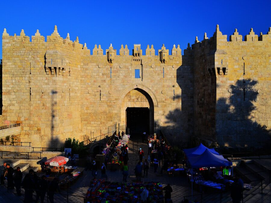 Damascus Gate in Jerusalem's northern Old City wall glowing gold in late-afternoon sun, its crenellated towers against a deep blue sky and market stalls in the plaza below