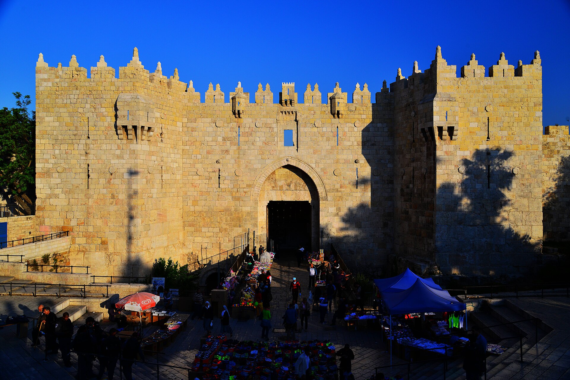 Damascus Gate in Jerusalem's northern Old City wall glowing gold in late-afternoon sun, its crenellated towers against a deep blue sky and market stalls in the plaza below