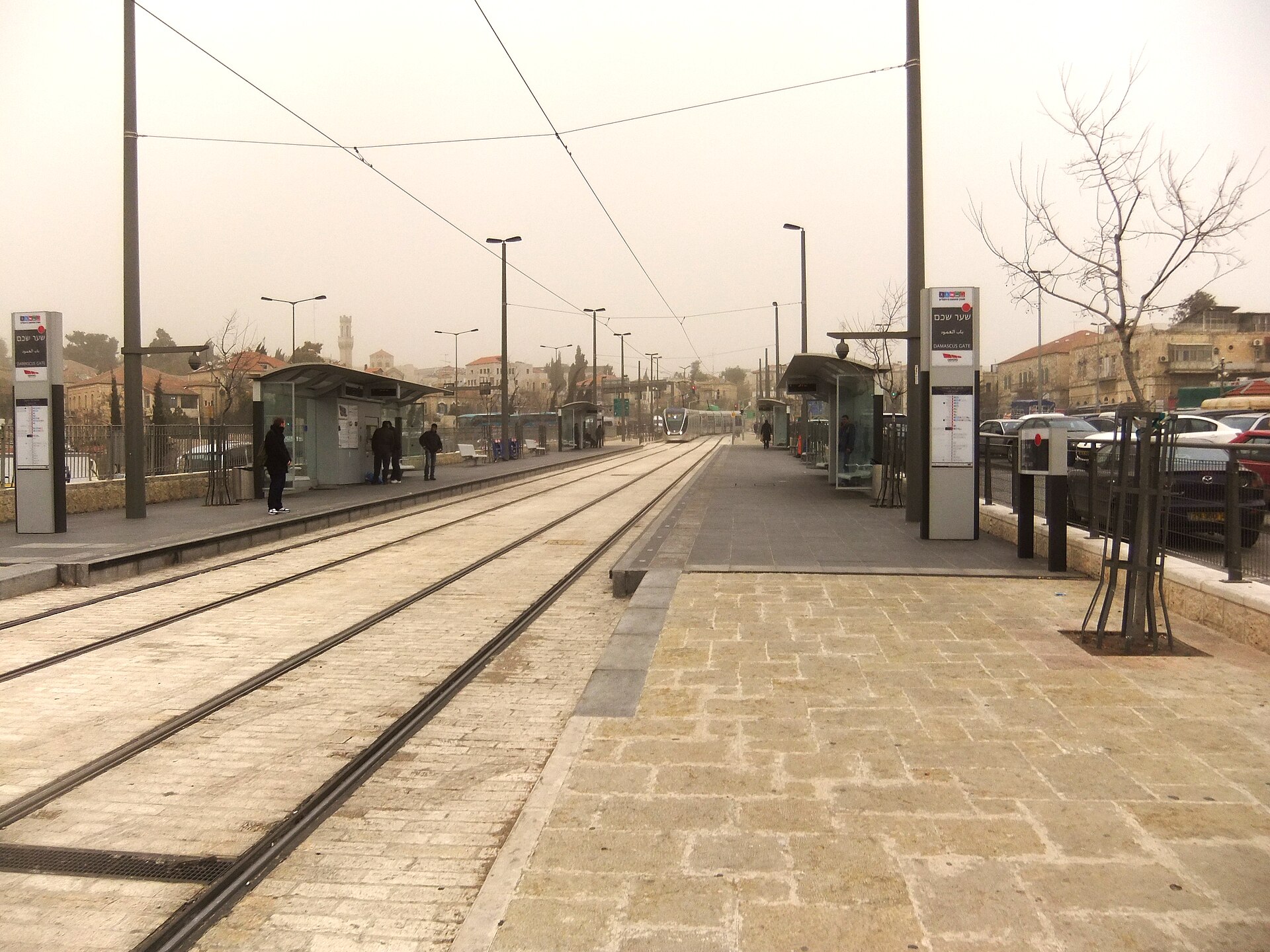 The Damascus Gate light rail stop in Jerusalem, with its trilingual Hebrew, Arabic and English sign, twin tracks and a tram approaching in hazy weather