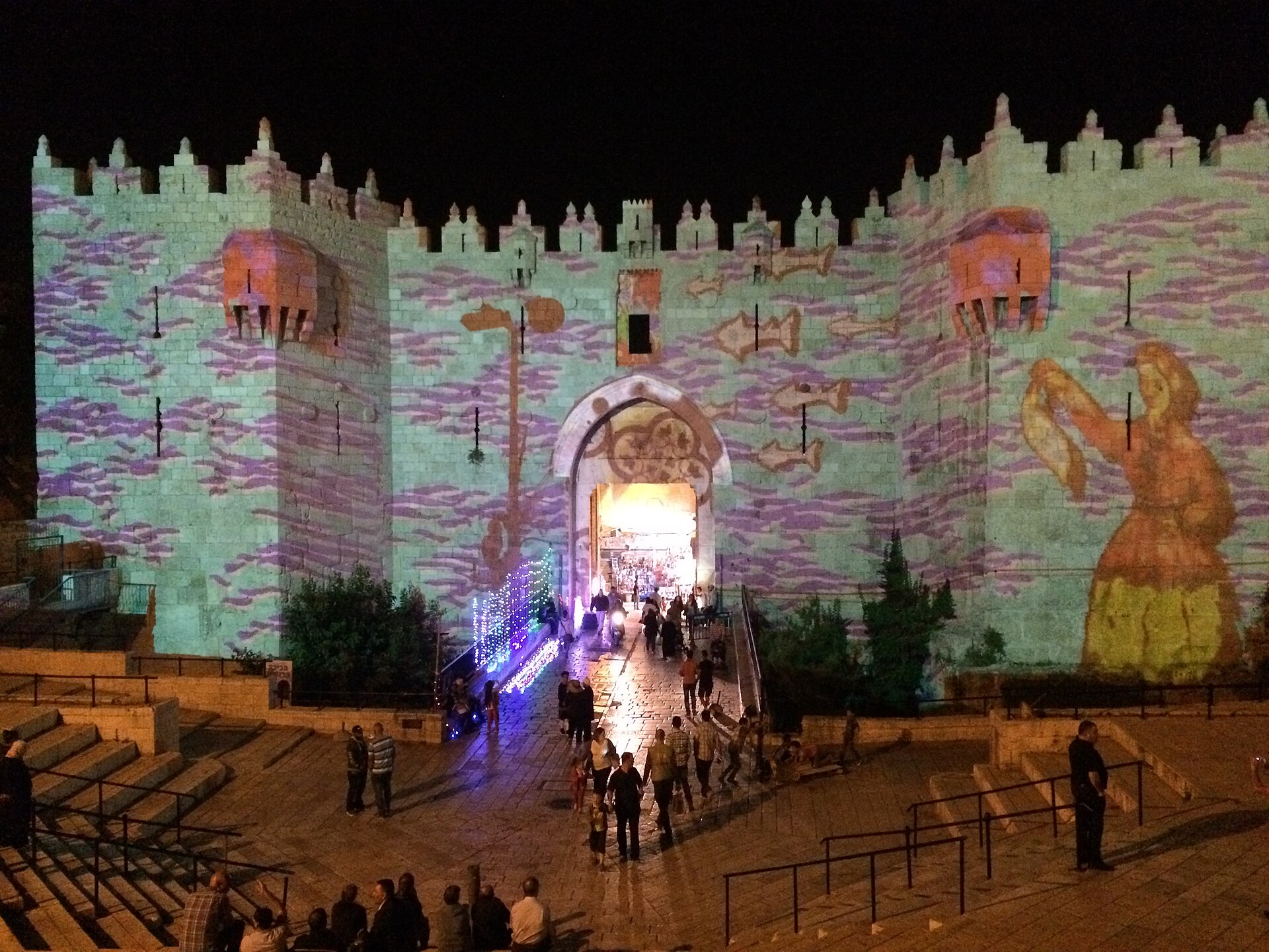 Damascus Gate at night covered in projected artwork of purple waves, fish and a painted female figure, with people walking across the plaza and sitting on the steps