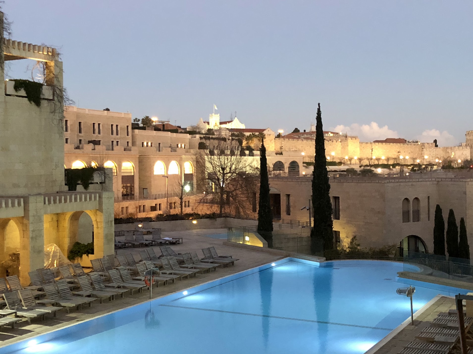 The David Citadel Hotel's lit swimming pool and sunloungers at dusk, with arcaded stone buildings and the floodlit Old City walls of Jerusalem beyond