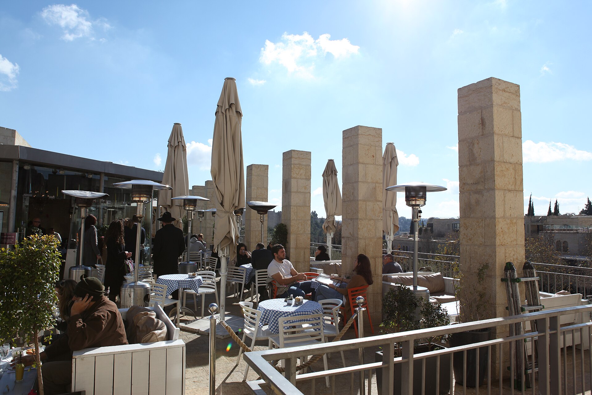 Diners at blue gingham tables on a sunny café terrace at Mamilla in Jerusalem, set among square stone pillars, furled parasols and patio heaters