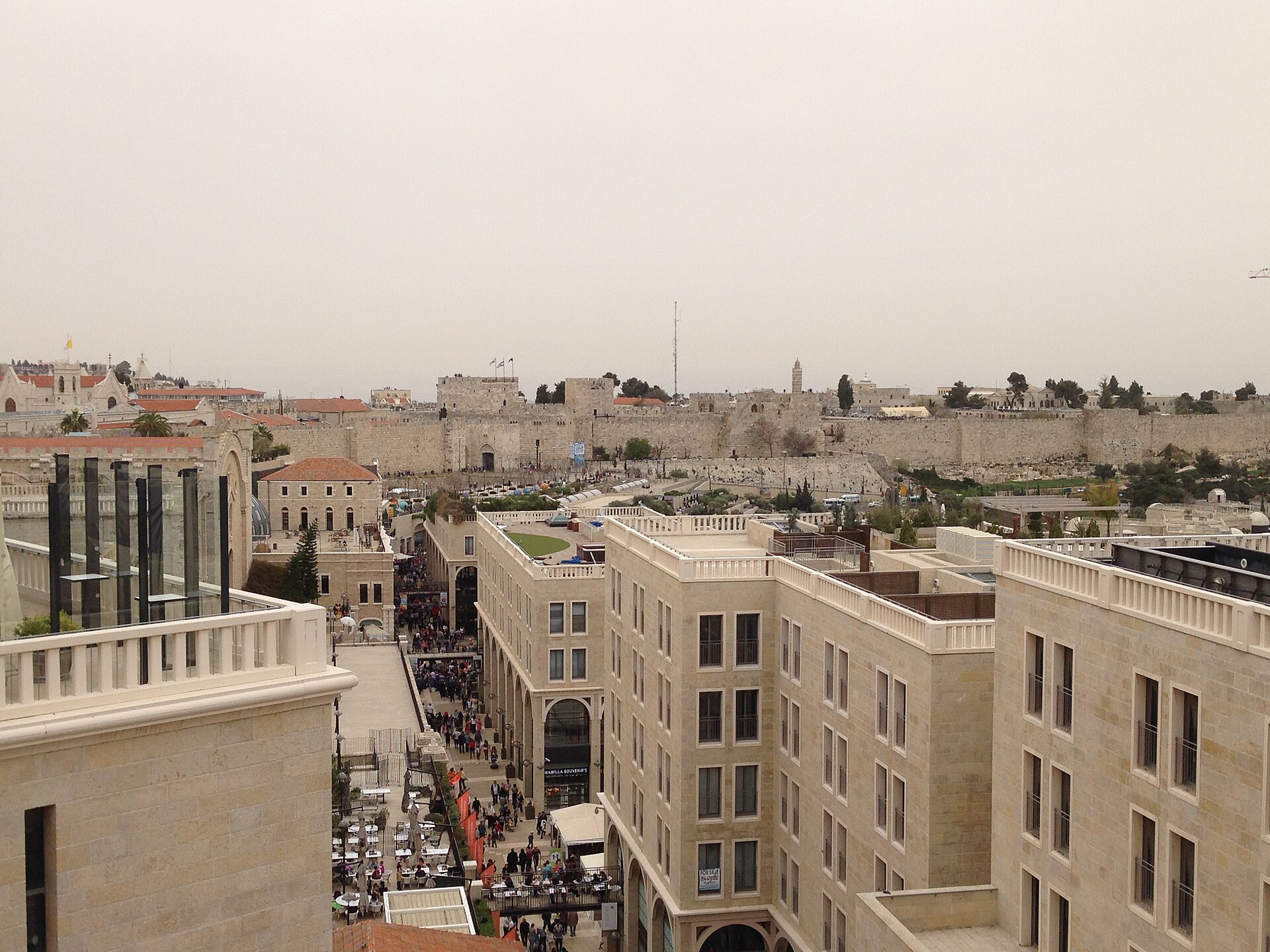 Elevated view down Mamilla Avenue in Jerusalem, crowded with shoppers and café tables between pale stone buildings, leading to Jaffa Gate and the Old City walls