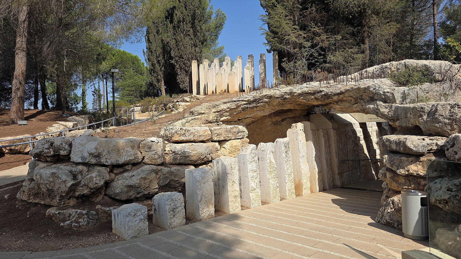 Entrance to Yad Vashem's Children's Memorial beneath a rock overhang, lined with white stone pillars of different heights, with taller broken pillars on the hill above