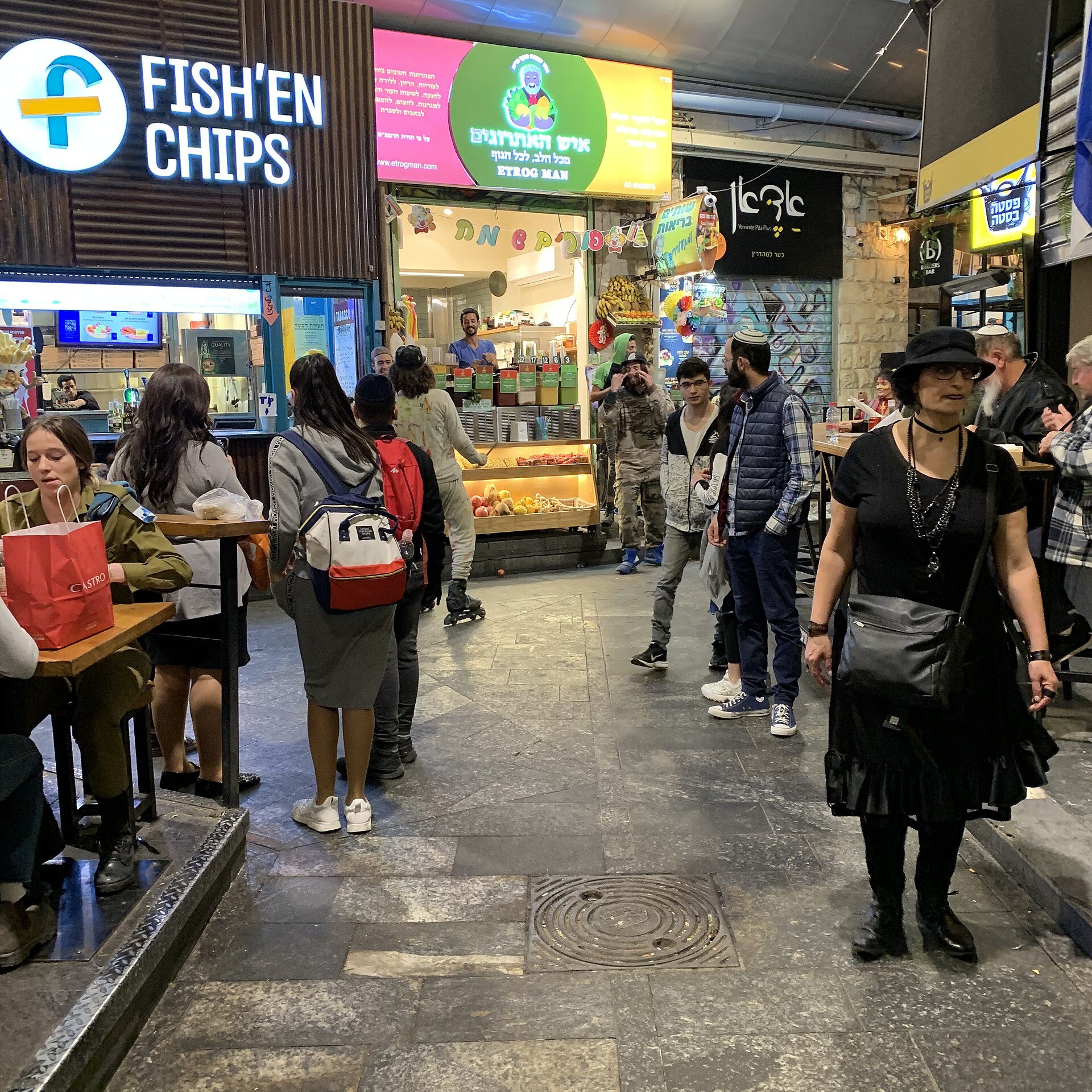 Evening crowd in a covered lane of Mahane Yehuda Market, with a Happy Purim banner and clown decorations above the Etrog Man juice bar and a fish and chips counter