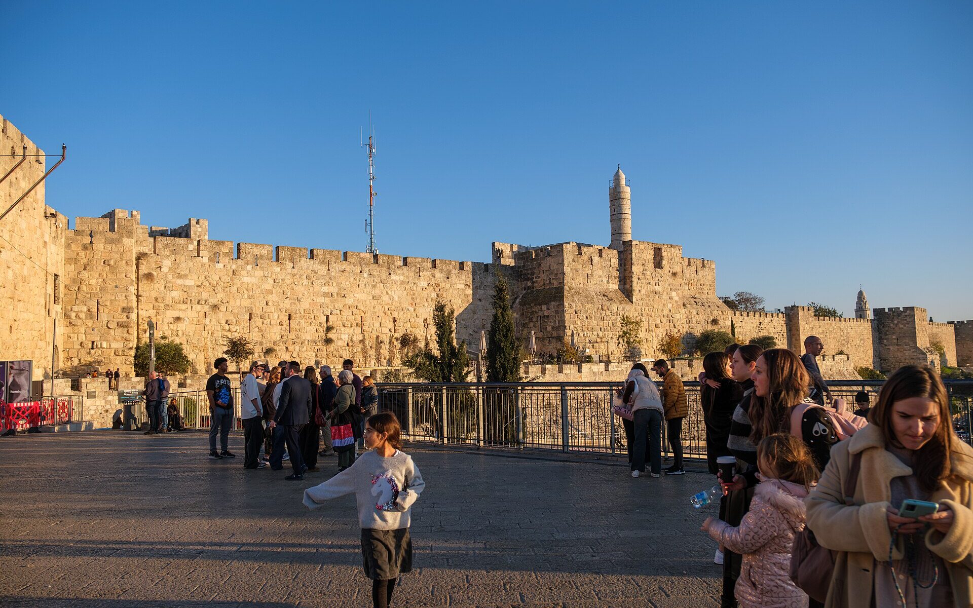Families stroll on the plaza outside Jaffa Gate in late-afternoon sun, a girl running in front, with the Old City walls and Tower of David minaret behind