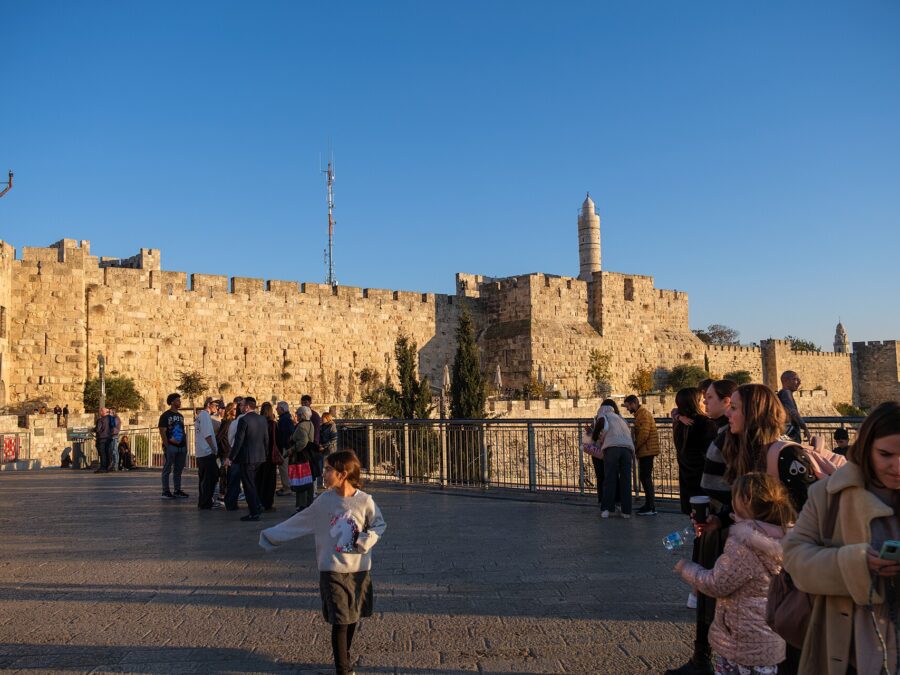 Families stroll on the plaza outside Jaffa Gate in late-afternoon sun, a girl running in front, with the Old City walls and Tower of David minaret behind