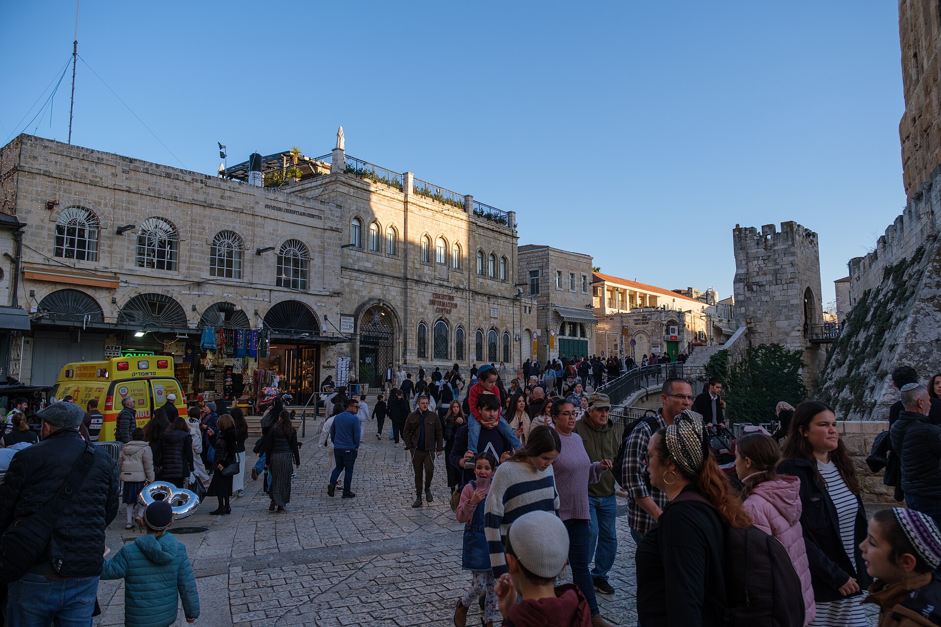 Families with young children crowd the square just inside Jaffa Gate, one child riding on a parent's shoulders, beside the stone wall of the Tower of David