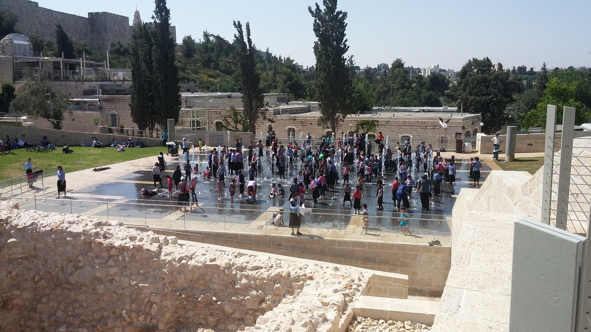 Families with young children play among the Teddy Park fountain jets, seen from above past a rough stone wall, with picnickers on the lawn and the Old City wall behind