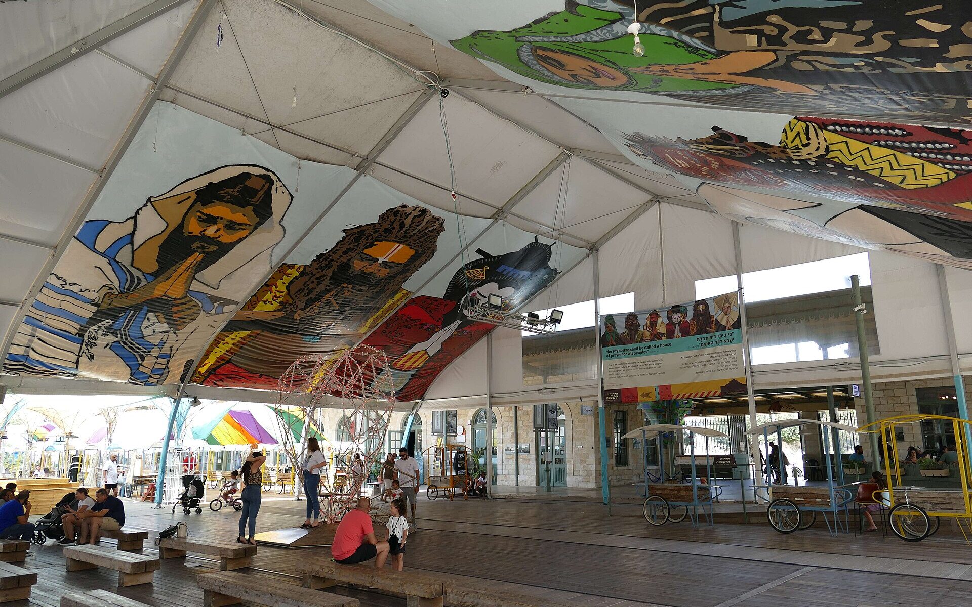 Families with young children relaxing on wooden benches under a large painted canopy at Jerusalem's First Station, beside the restored stone station building