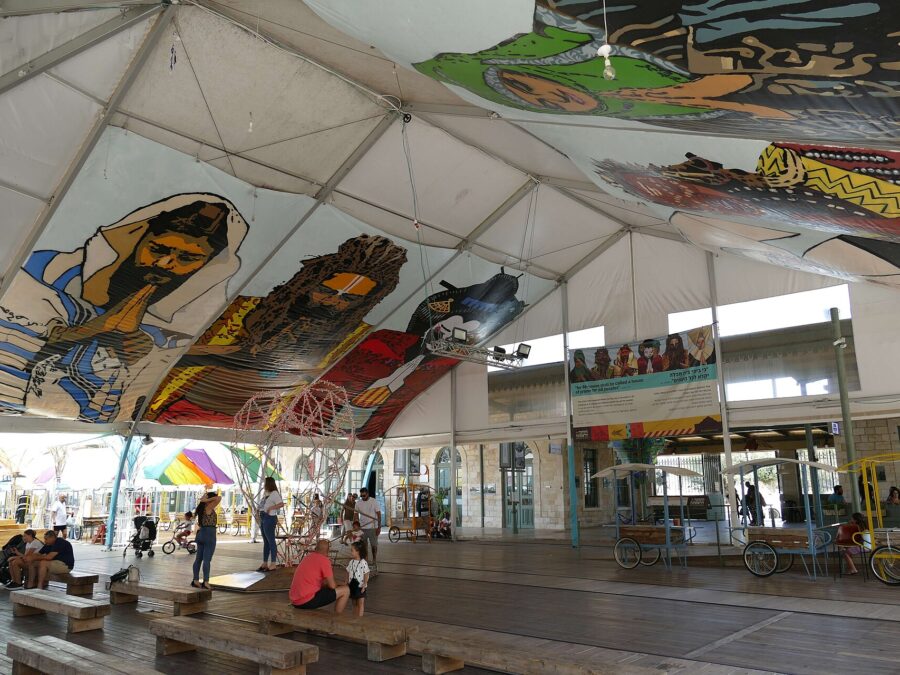 Families with young children relaxing on wooden benches under a large painted canopy at Jerusalem's First Station, beside the restored stone station building
