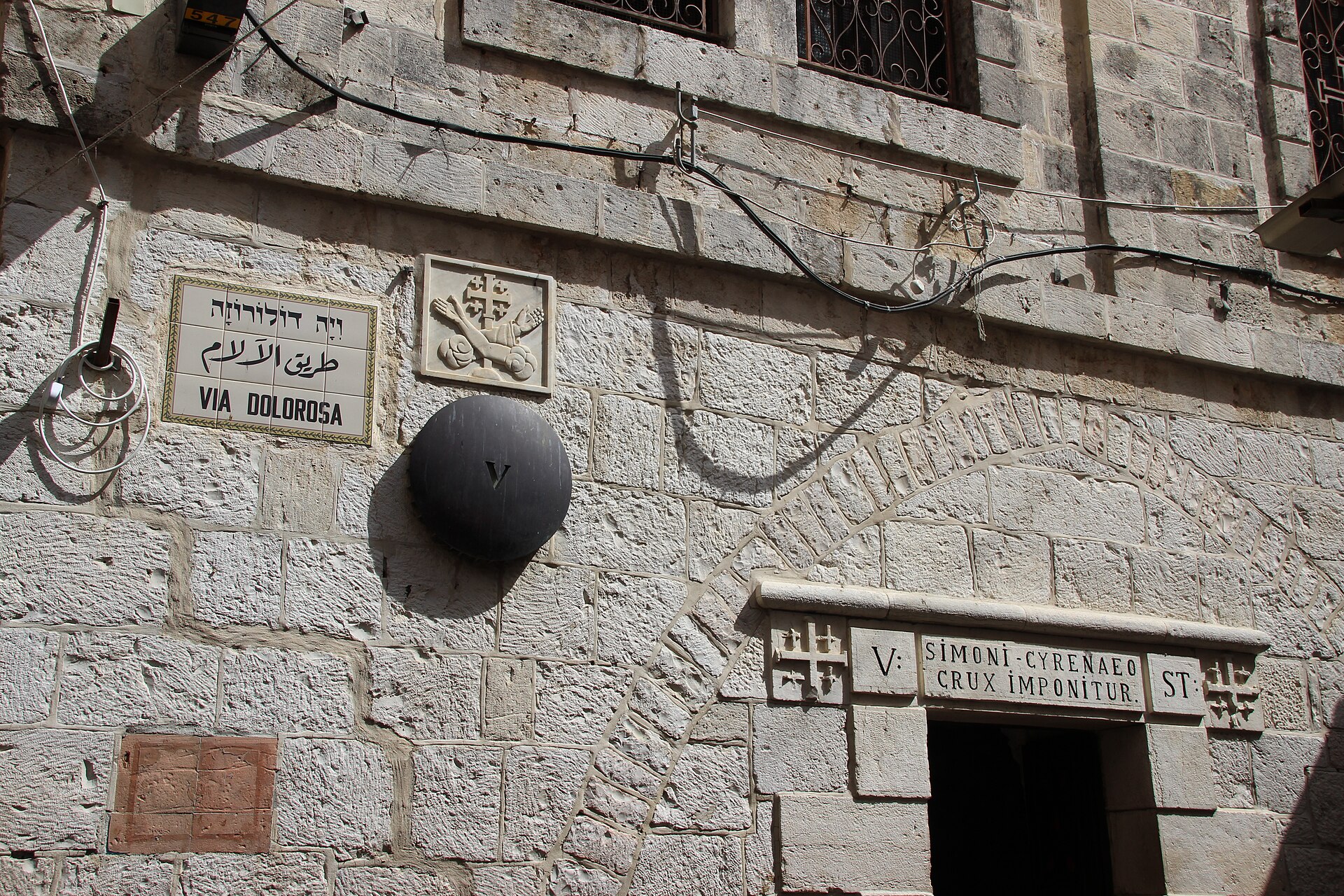 Fifth Station of the Cross on the Via Dolorosa, with a round metal marker bearing the numeral V, a trilingual street sign and a Latin inscription above a doorway