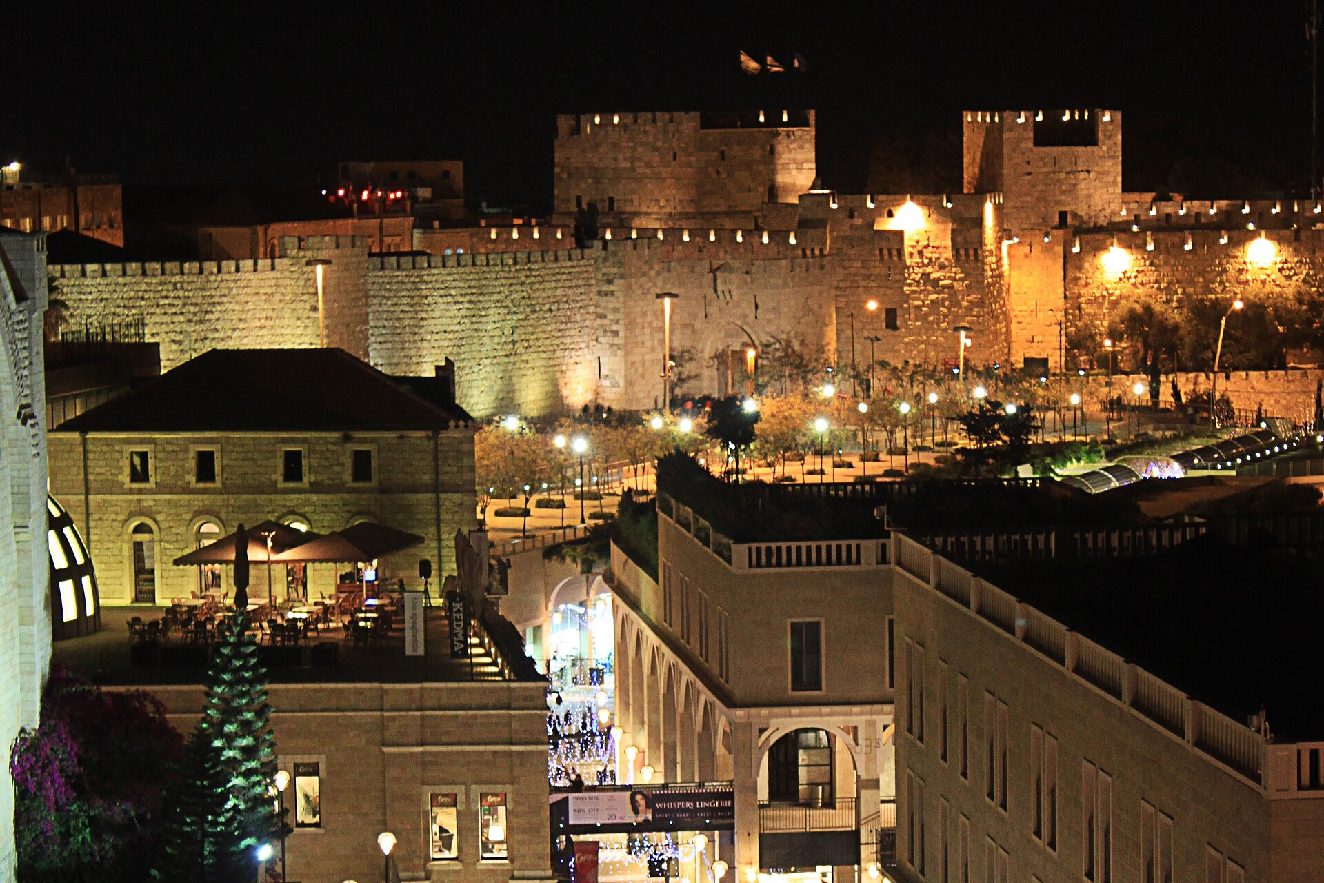 Floodlit Old City walls and Jaffa Gate at night above Mamilla Avenue strung with fairy lights, seen from a rooftop restaurant in Jerusalem