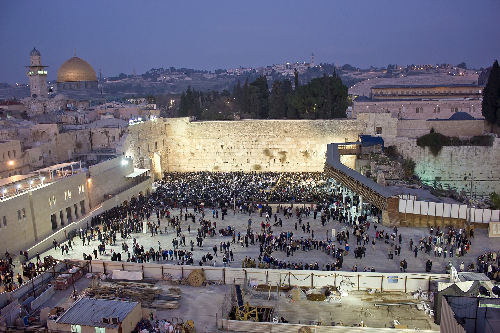 The floodlit Western Wall at dusk on a Friday evening, its plaza filling with worshippers for Shabbat prayers, with the Dome of the Rock and Mount of Olives beyond