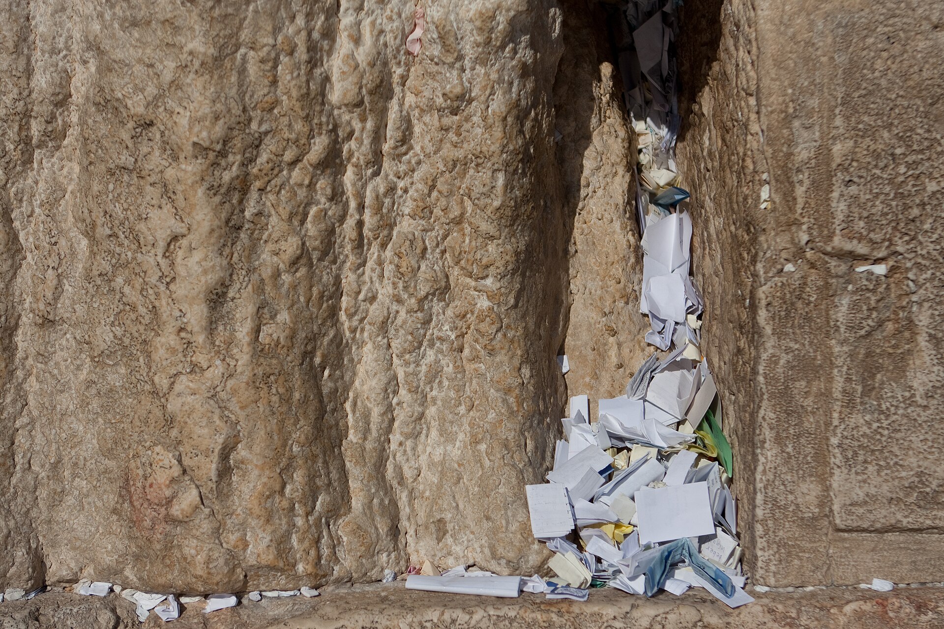 Folded paper prayer notes packed into a vertical crack between two massive weathered stones of the Western Wall, spilling into a heap at its base