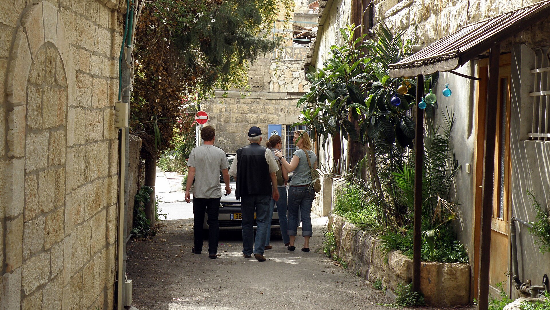 Four visitors seen from behind stroll down a narrow stone-walled lane in Ein Karem, past leafy plants and coloured glass baubles hanging from a small awning