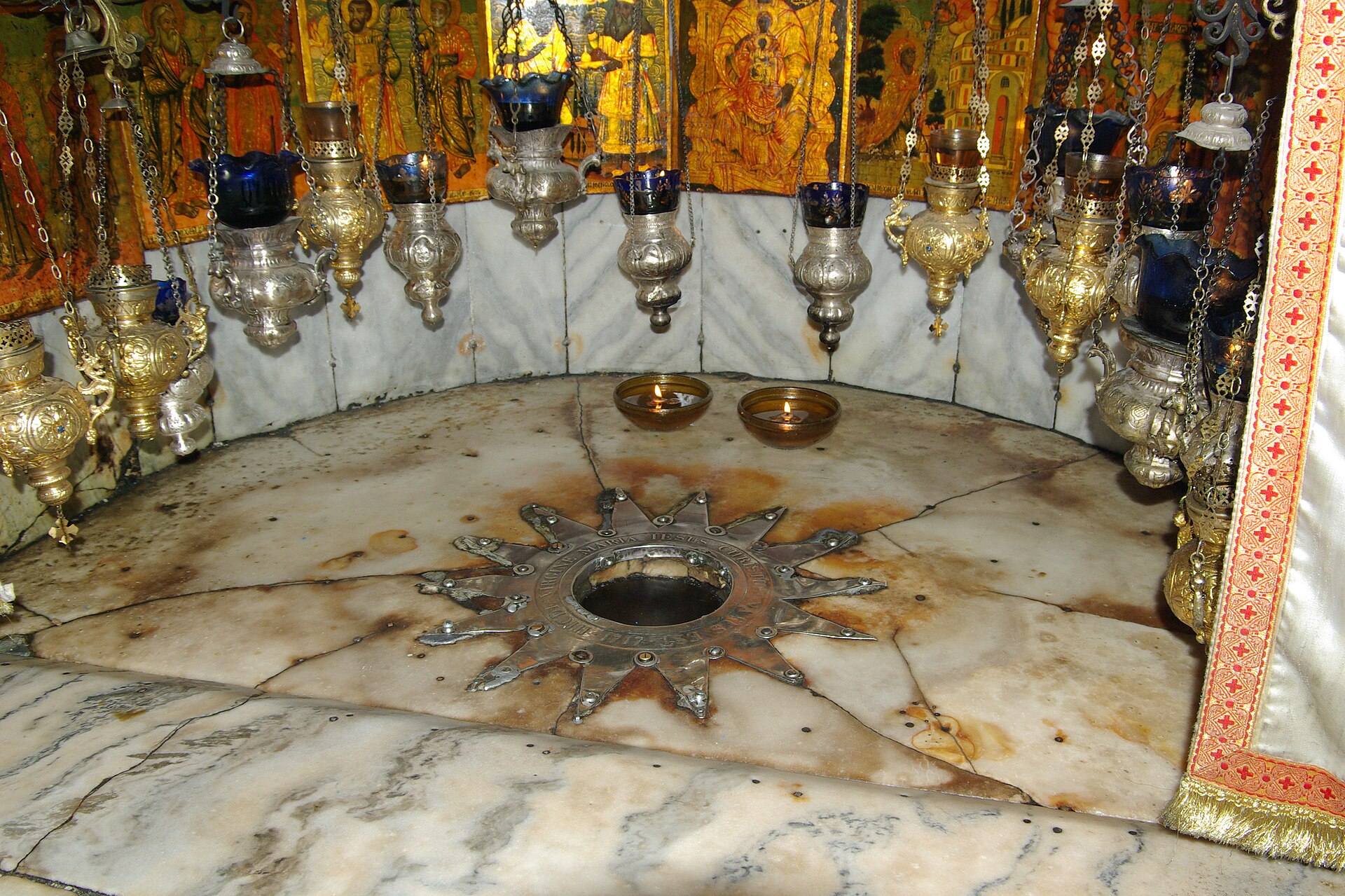 Fourteen-pointed silver star set in the marble floor of a niche in the Grotto of the Nativity, Bethlehem, beneath rows of hanging silver oil lamps and icons