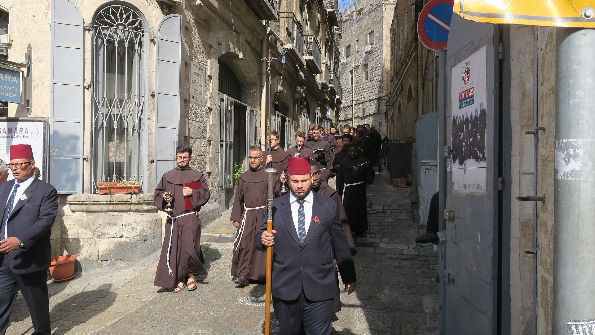 Franciscan friars in brown habits walk in procession along a narrow Christian Quarter lane in Jerusalem, led by kawas in red fezzes carrying ceremonial staffs