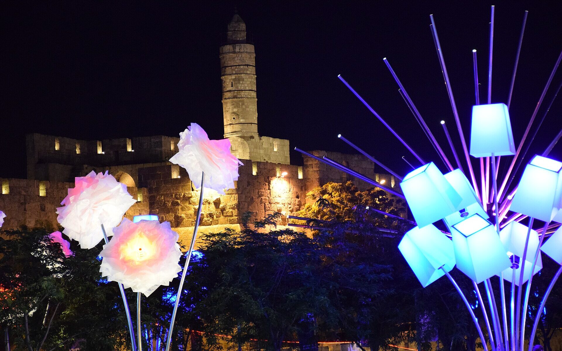Glowing flower and lampshade light sculptures in front of the floodlit Tower of David citadel and its minaret at night during the Jerusalem Festival of Light