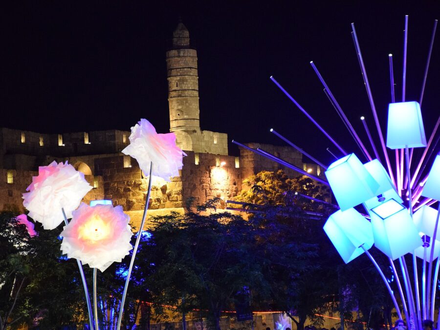 Glowing flower and lampshade light sculptures in front of the floodlit Tower of David citadel and its minaret at night during the Jerusalem Festival of Light