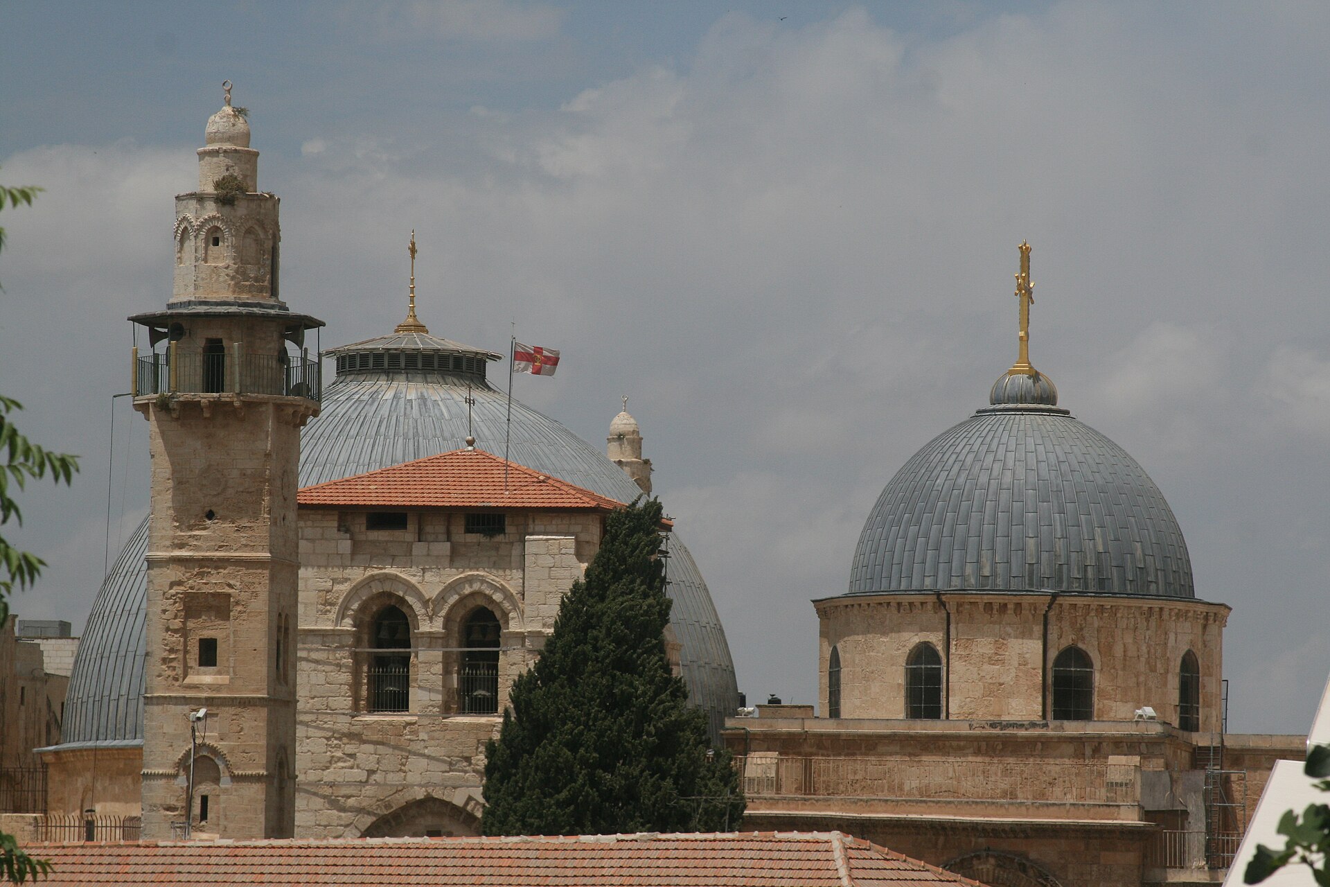 Grey domes and the stone bell tower of the Church of the Holy Sepulchre rising above tiled rooftops in Jerusalem's Christian Quarter, beside the Mosque of Omar minaret