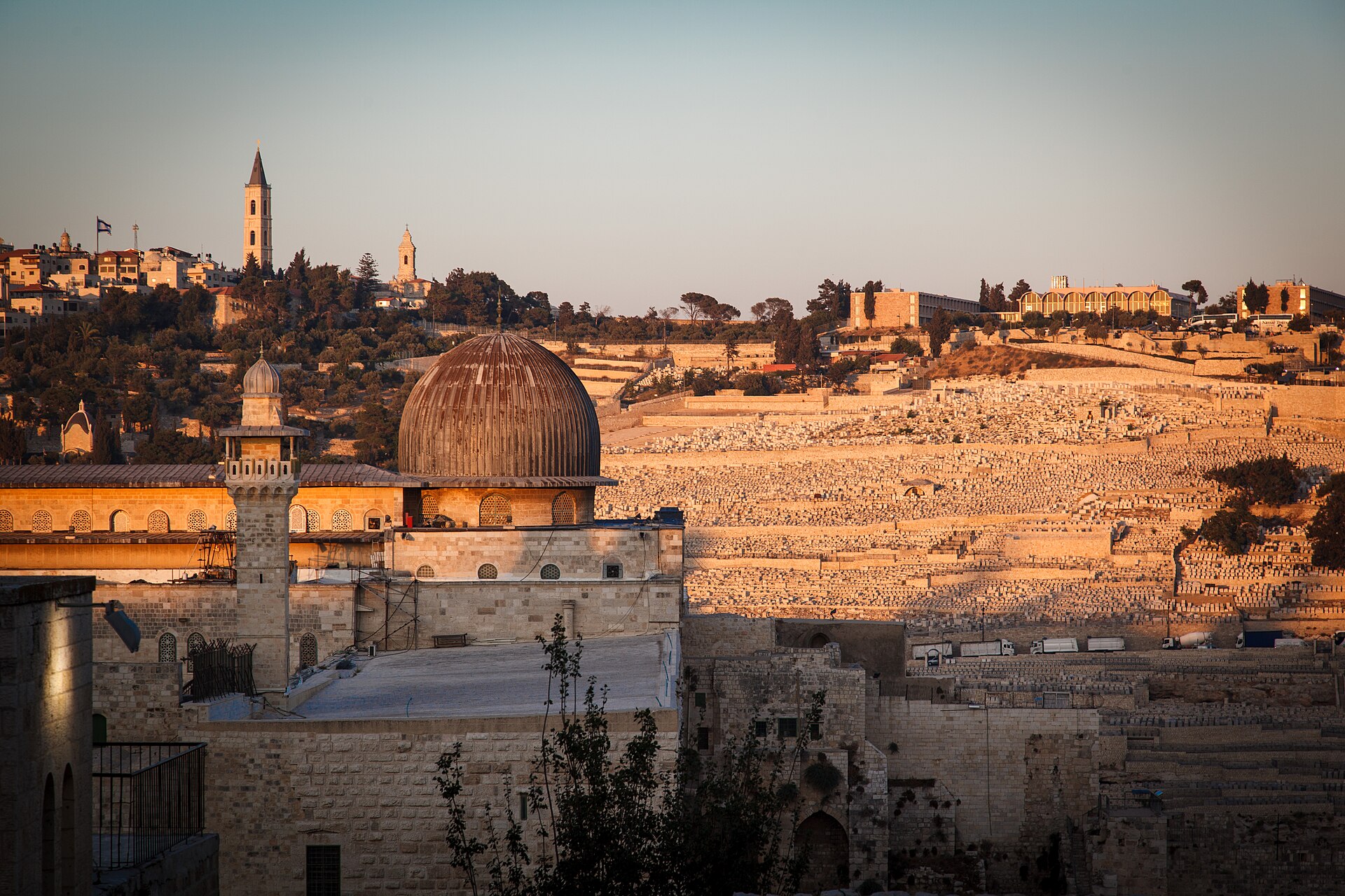 Grey ribbed dome of the Al-Aqsa Mosque and a square stone minaret in low evening sun, with the Mount of Olives and its hillside cemetery rising behind