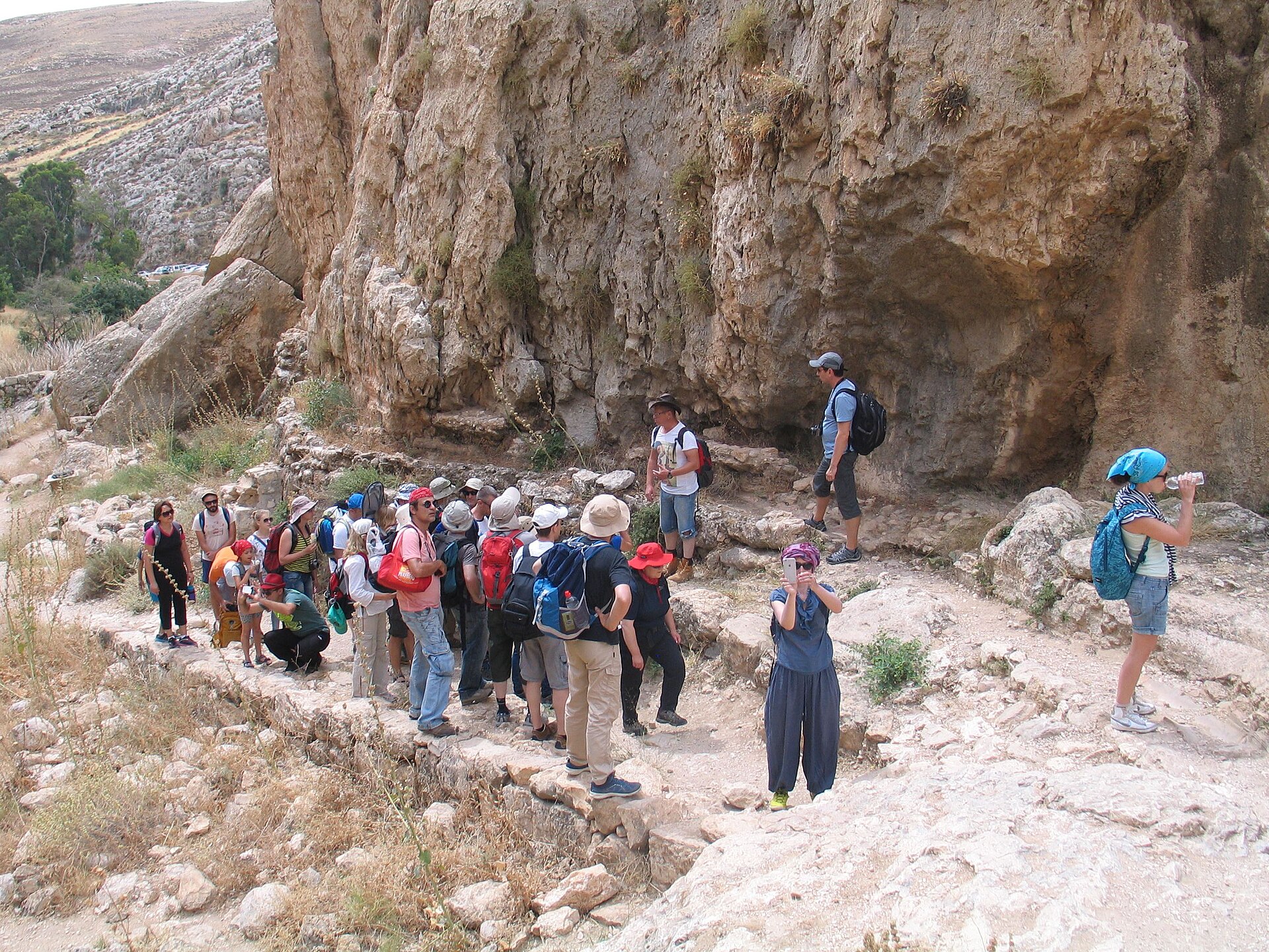 Group of hikers with sun hats and backpacks on a rocky trail below an overhanging limestone cliff in Nahal Prat, the upper part of Wadi Qelt
