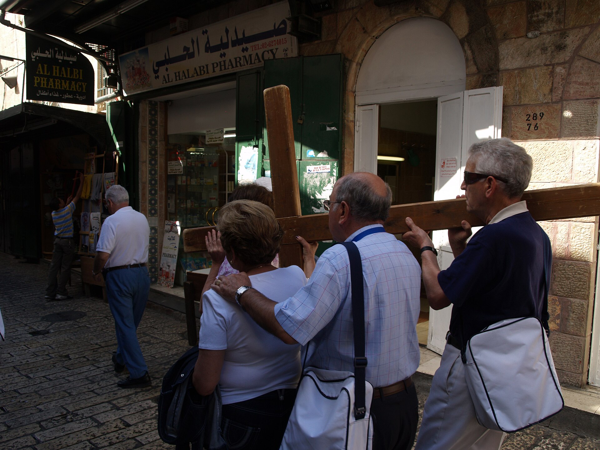 Group of pilgrims shouldering a heavy wooden cross along a stone-paved lane of the Via Dolorosa, walking past shopfronts in Jerusalem's Old City