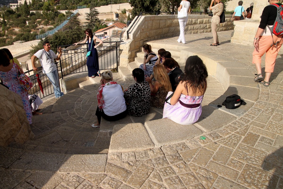 A guide gestures as he talks to a small group of women seated on curved stone steps at a viewpoint on the Mount of Olives in Jerusalem