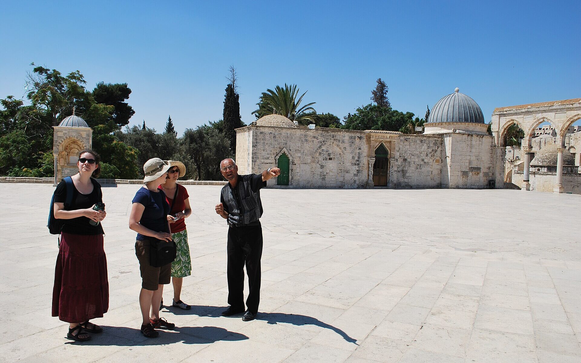 A guide points out buildings to three visitors on the sunlit stone esplanade of the Temple Mount (Haram al-Sharif), with a domed medieval building and arches behind