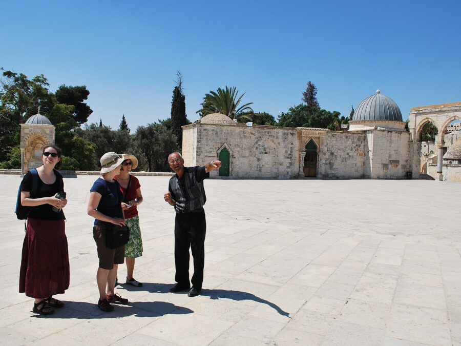 A guide points out buildings to three visitors on the sunlit stone esplanade of the Temple Mount (Haram al-Sharif), with a domed medieval building and arches behind