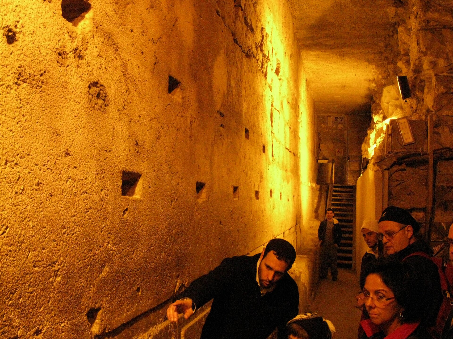 A guide points out the huge Herodian stones to a small tour group in a narrow, lamp-lit passage of the Western Wall Tunnels, with stairs behind