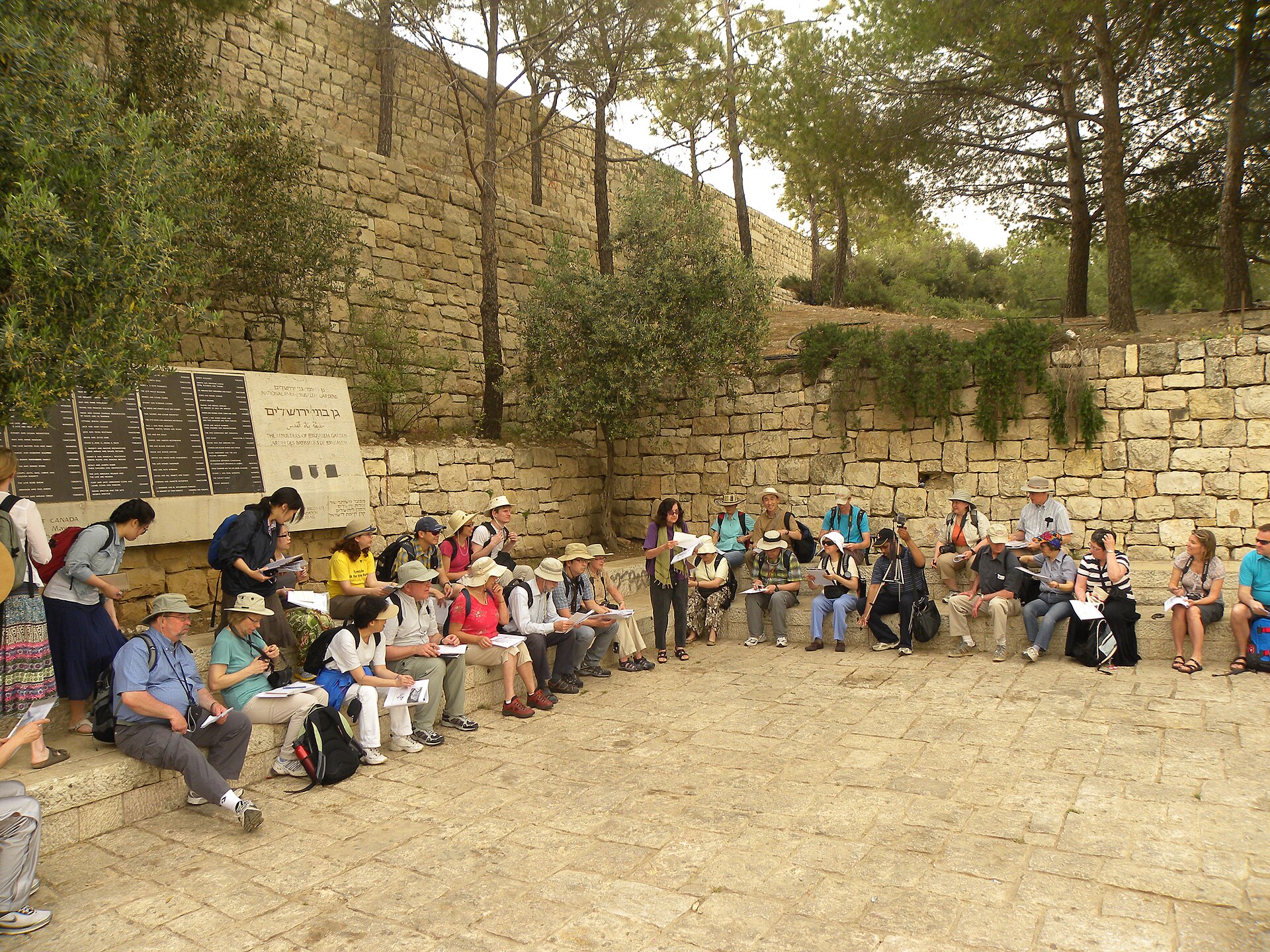 A guide reads from her notes to a tour group seated on stone benches in a shaded garden below stone walls, just outside the south-west corner of Jerusalem's Old City
