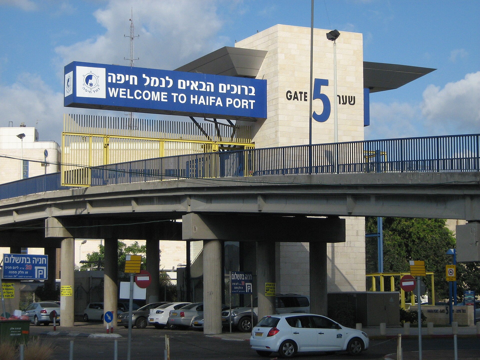 The Gate 5 entrance to Haifa Port with a blue Welcome to Haifa Port sign in Hebrew and English, a pedestrian bridge and parked cars