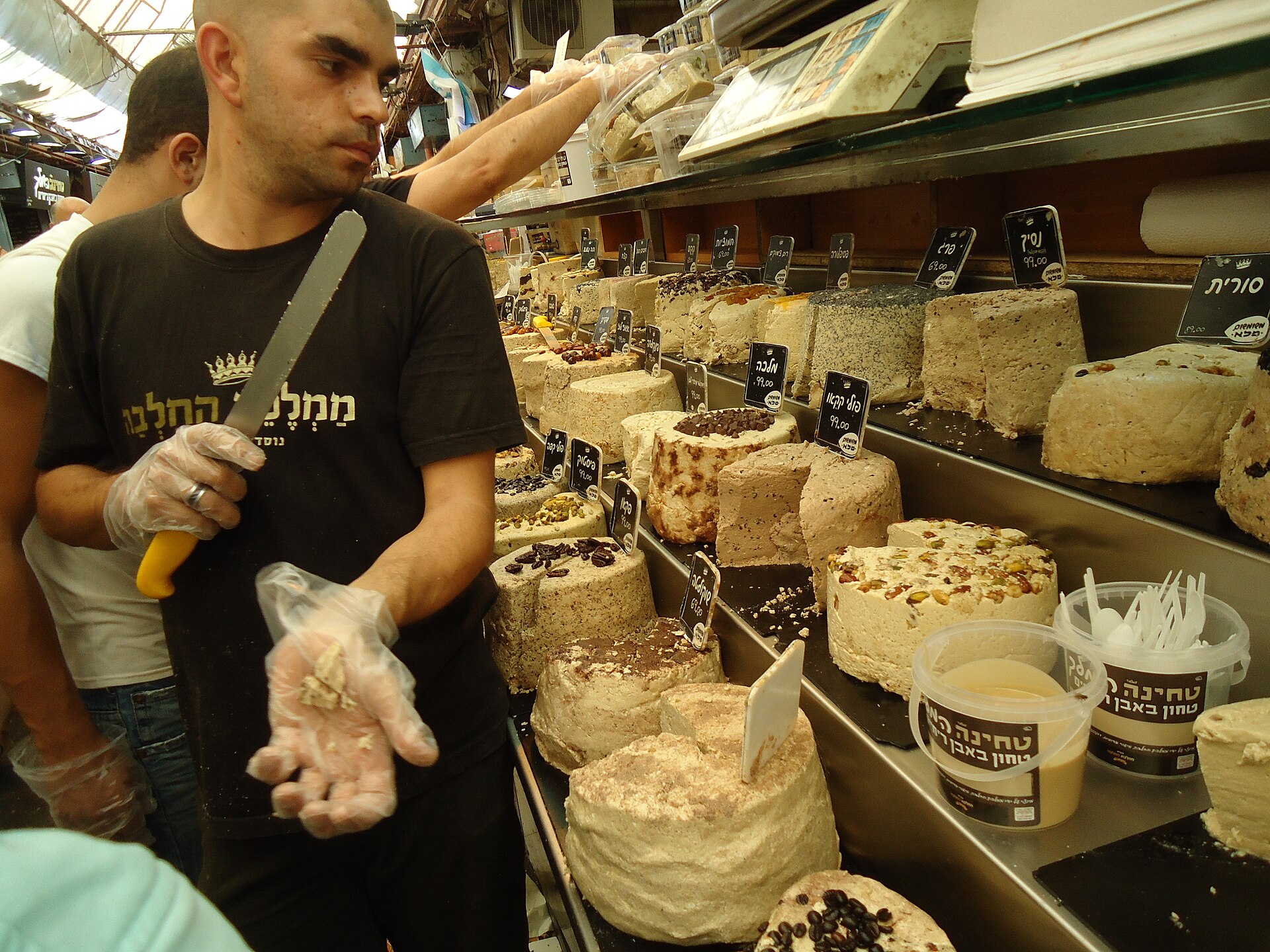 A Halva Kingdom vendor in a crown-logo shirt holds a knife beside wheels of halva in many flavours with Hebrew price tags and tubs of stone-ground tahini