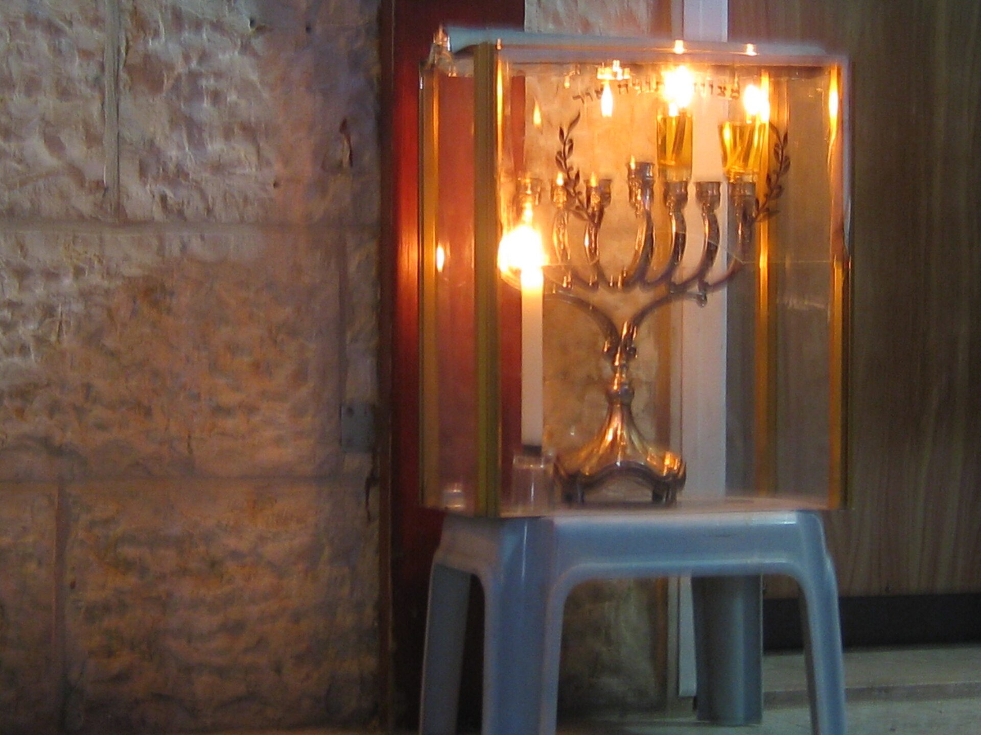 A lit silver hanukkiah inside a glass case standing on a stool outside a front door against a Jerusalem stone wall