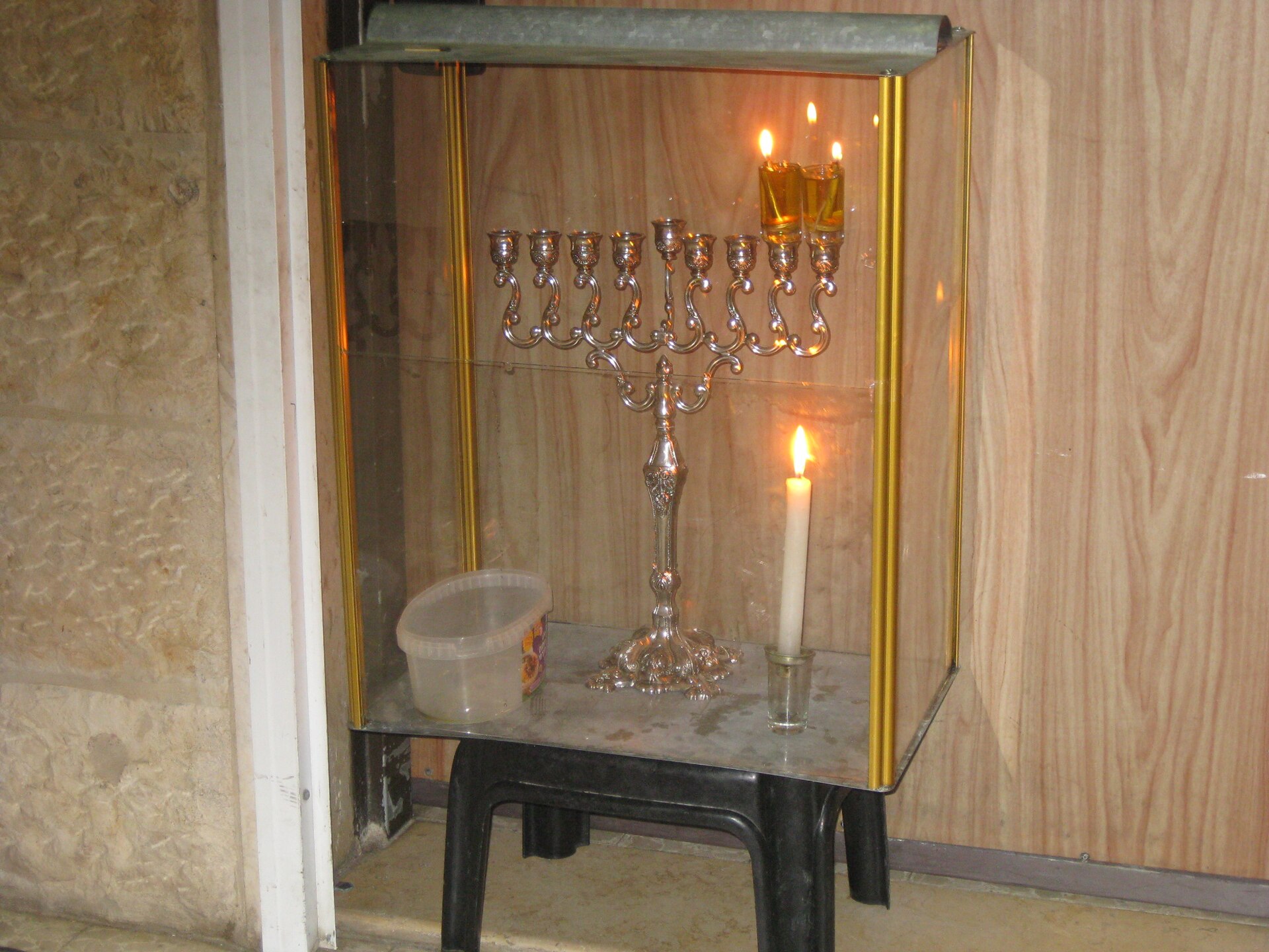 A hanukkiah with olive oil glasses lit inside a glass case beside a doorway, with a shammash candle standing next to it