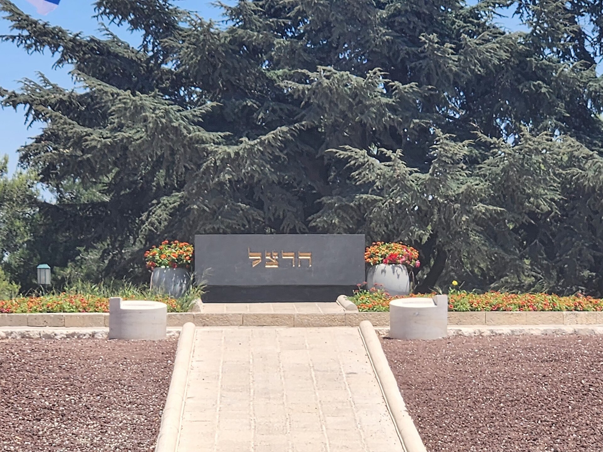 Theodor Herzl's black stone tomb on Mount Herzl, inscribed with his surname in gold Hebrew letters, between two planters of flowers with cedar trees behind
