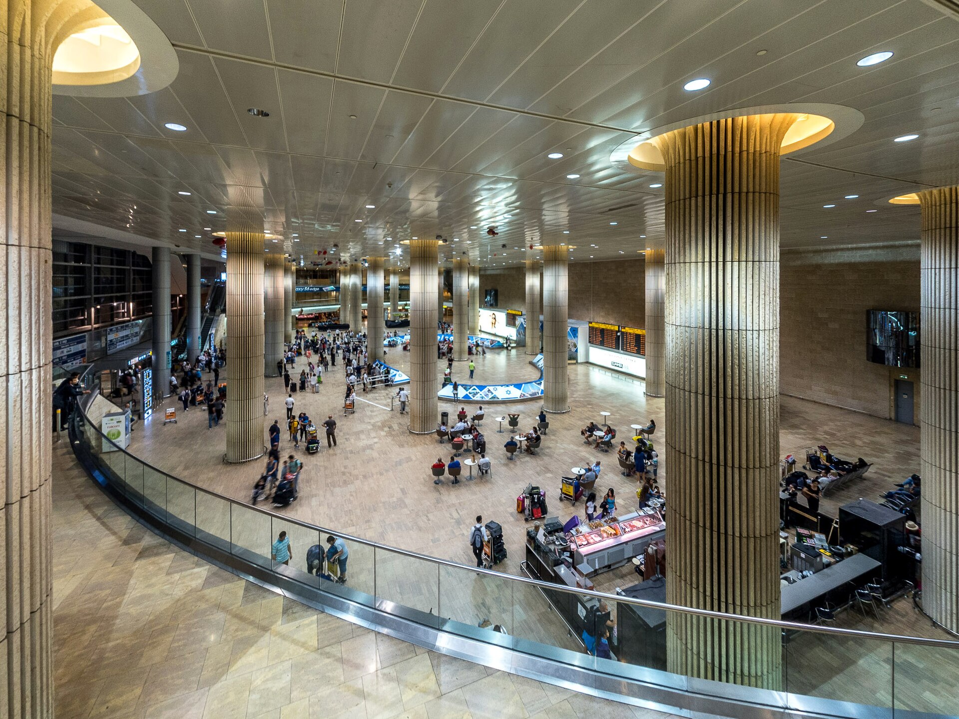 High view over the arrivals greeting hall of Ben Gurion Airport Terminal 3, with tall fluted columns, a café, flight boards and travellers pushing luggage trolleys